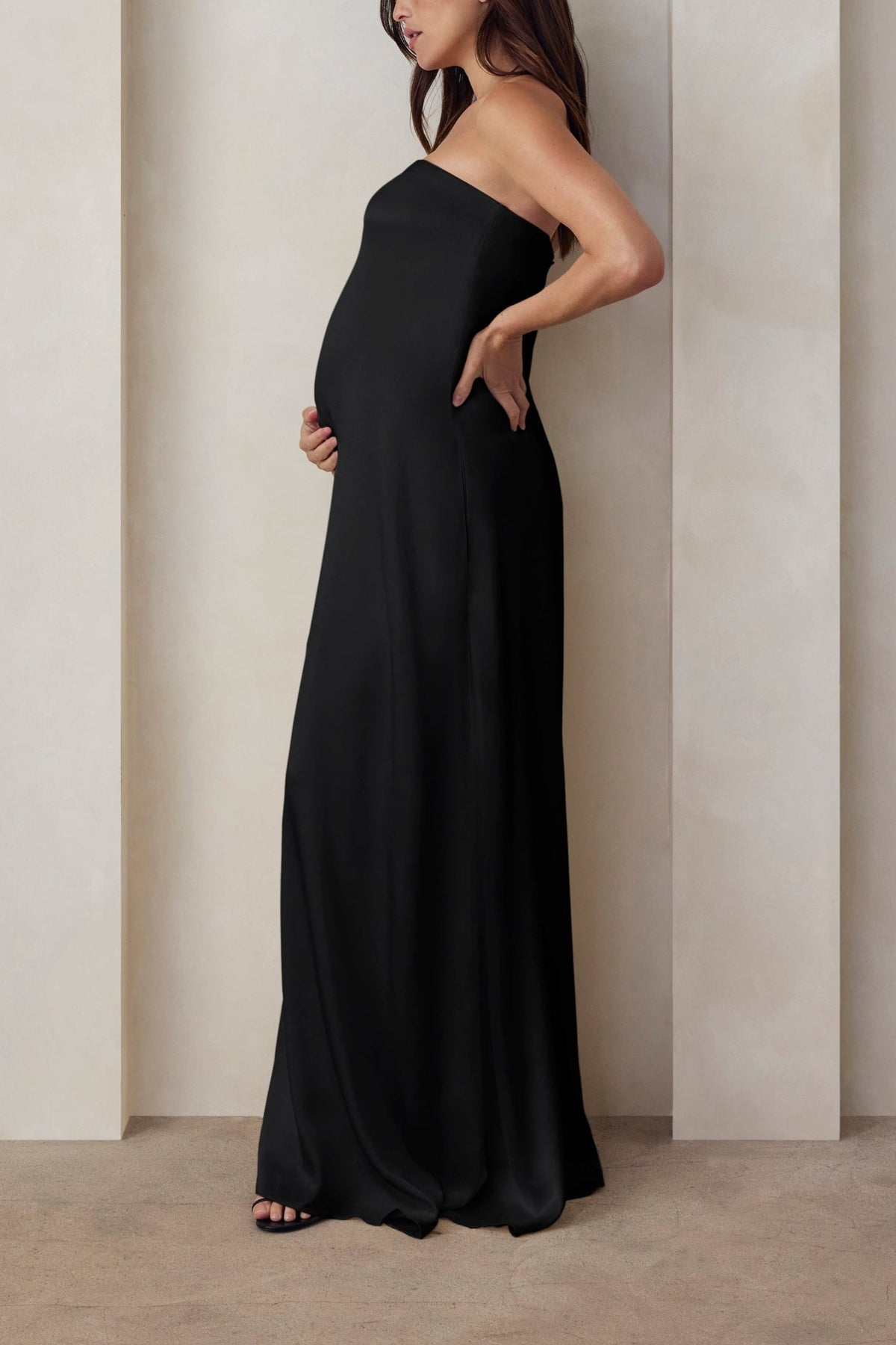 A pregnant woman wearing the Bumpsuit brushed satin strapless maxi maternity gown in black against a beige feature wall