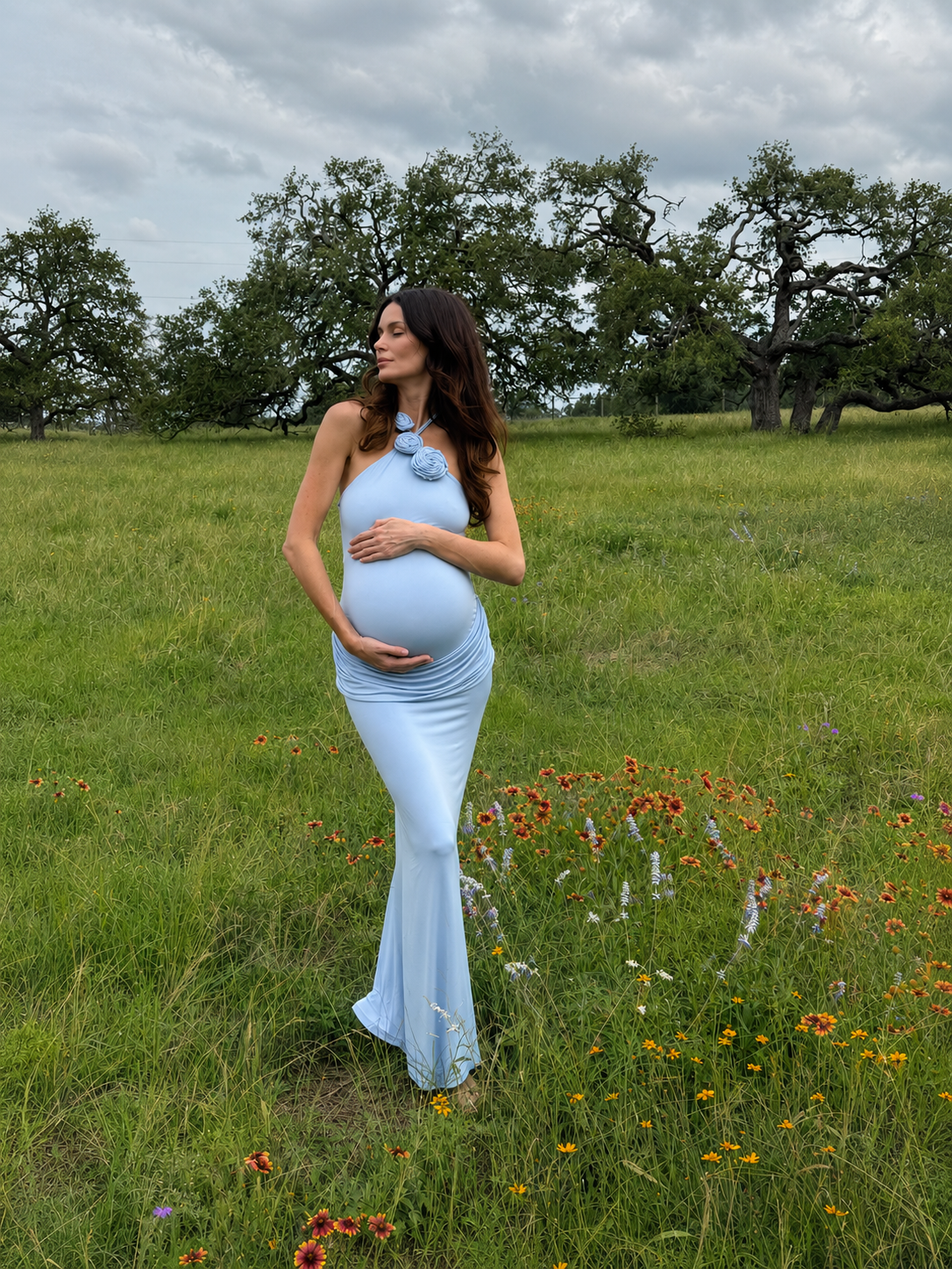 A pregnant woman stands in a grassy field with wildflowers and trees under a cloudy sky, wearing a light blue sleeveless maternity dress with a halter-style neckline and a large floral embellishment at the neck.