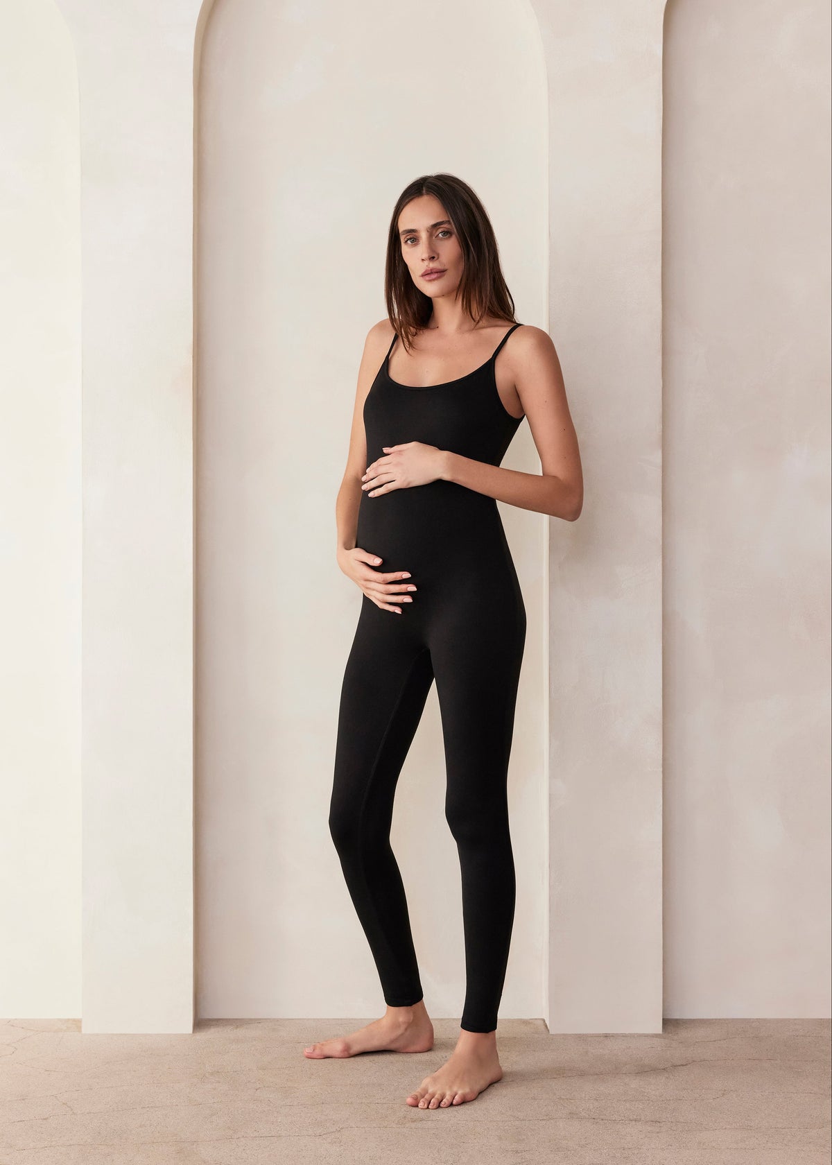 Woman wearing a Bumpsuit black sleeveless jumpsuit called the Kate  standing in a minimalistic setting.