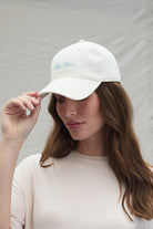 Woman wearing a white cap with baby mama written in blue txt and bumpsuit t-shirt against a plain background