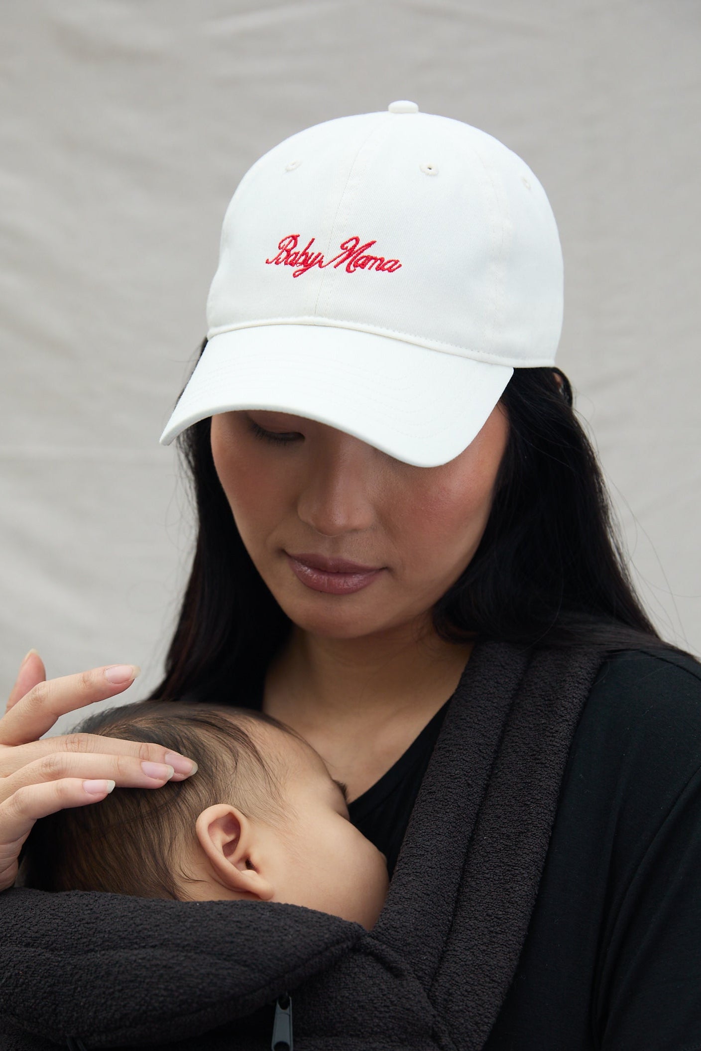 Woman wearing a white cap with 'Baby mama' text, holding a baby in a armadillo baby carrier against a neutral background.