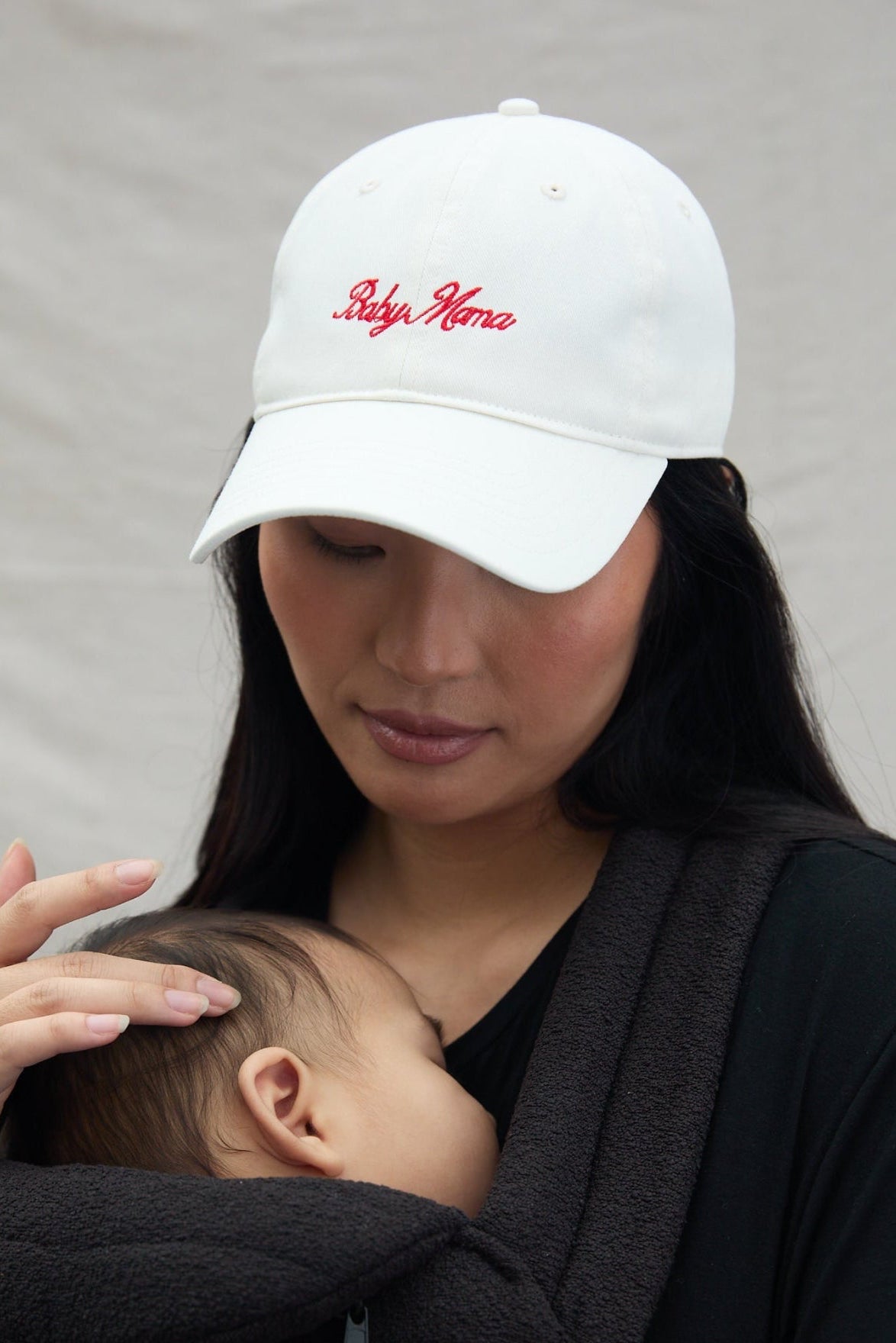 Woman wearing a white cap with 'Baby mama' text, holding a baby in a armadillo baby carrier against a neutral background.