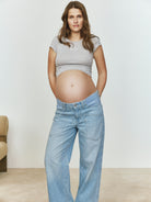 Pregnant woman wearing a heather gray baby mama tee and light blue jeans against a plain background
