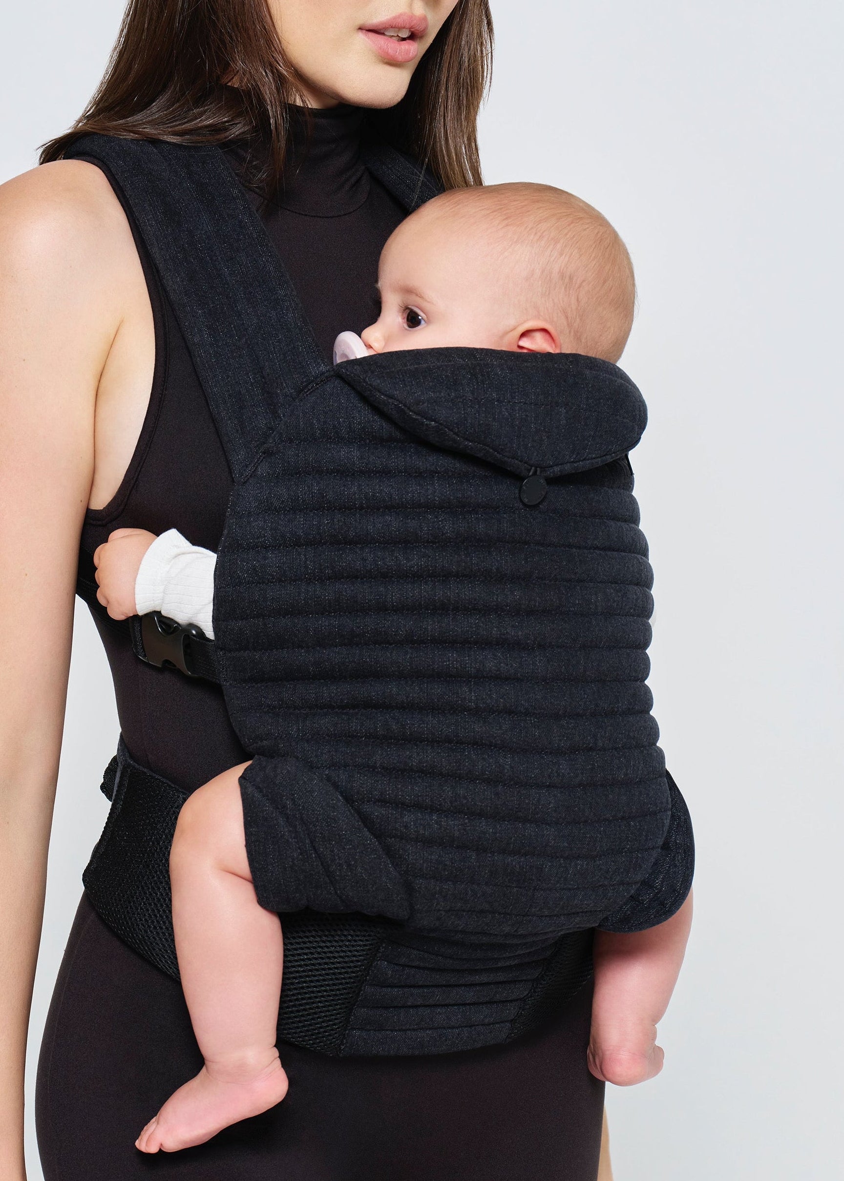 Woman holding a baby in a black denim baby carrier against a plain background