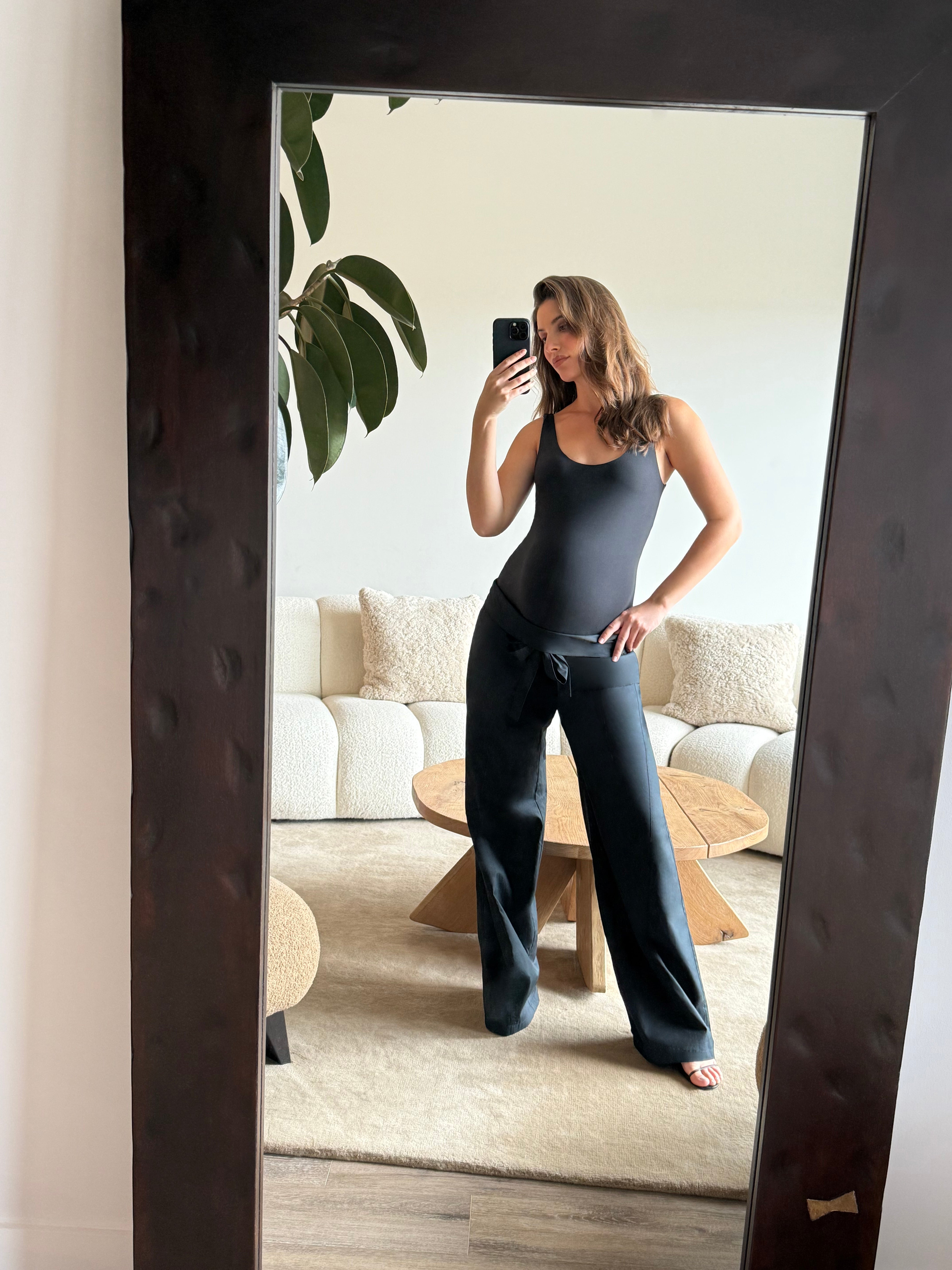 Pregnant women taking a mirror selfie in a modern living room wearing Bumpsuits tank bodysuit and jeans