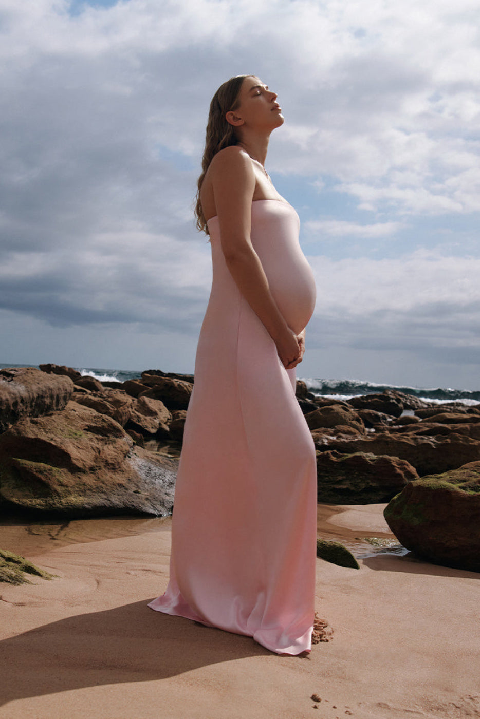A Pregnant woman wearing the Bumpsuit brushed satin gown in dusty pink on a beach.