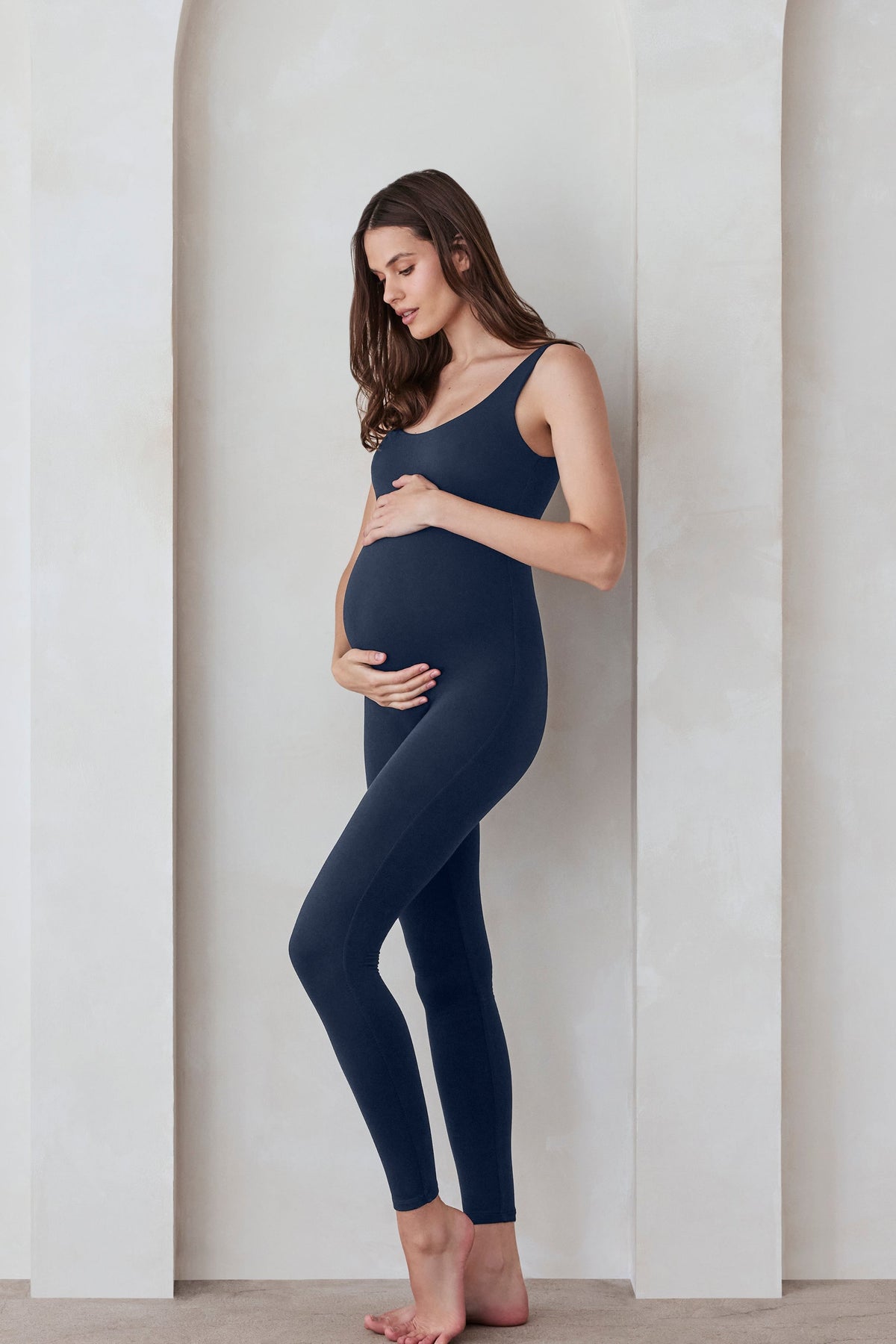 Pregnant woman wearing the Bumpsuit Lucy maternity scoop neck unitard in Navy blue standing against a feature wall