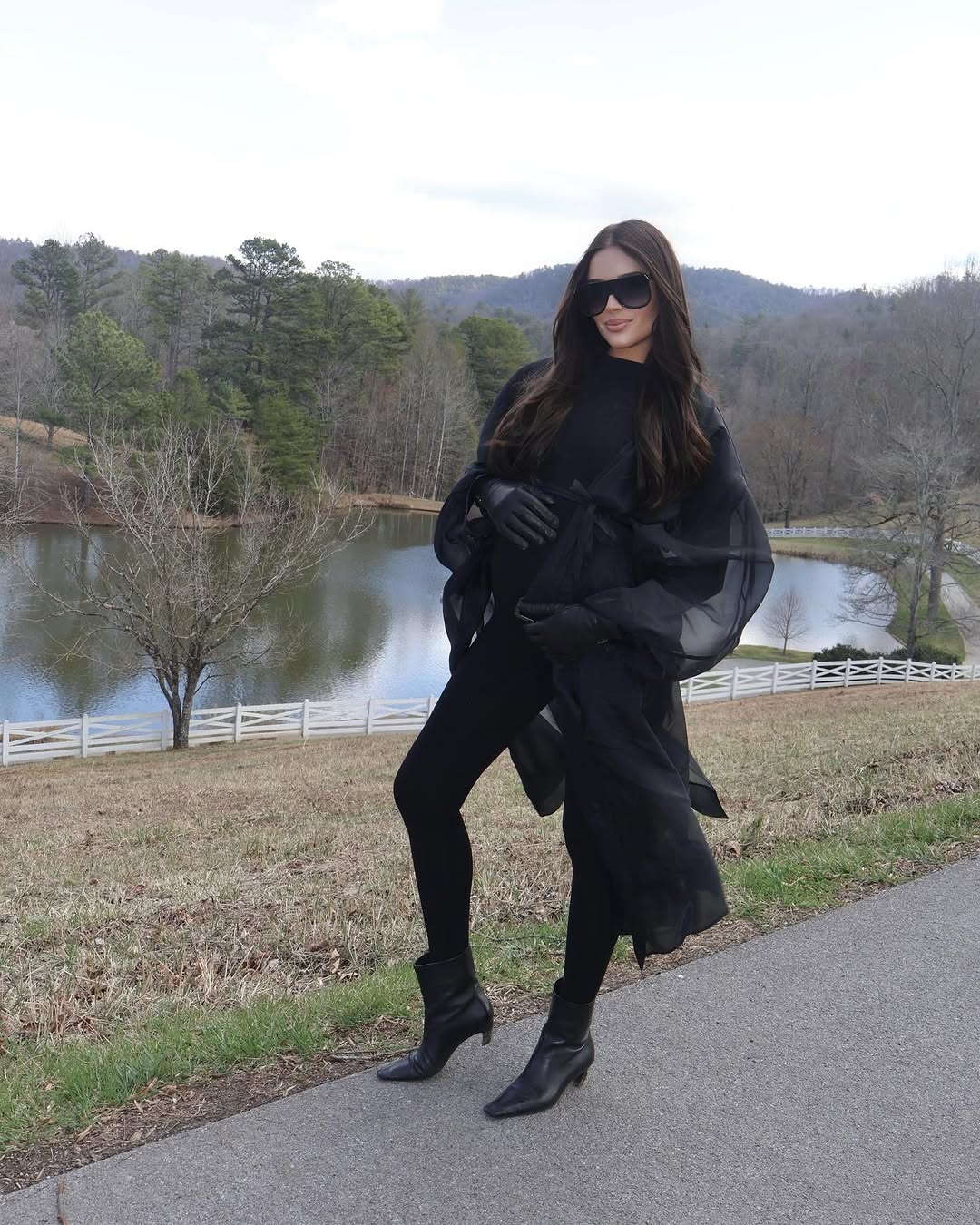 A woman posing outdoors in a black ensemble, featuring a black turtleneck jumpsuit by Bumpsuit, black gloves, and black ankle boots, set against a scenic background of a lake and wooded hills.