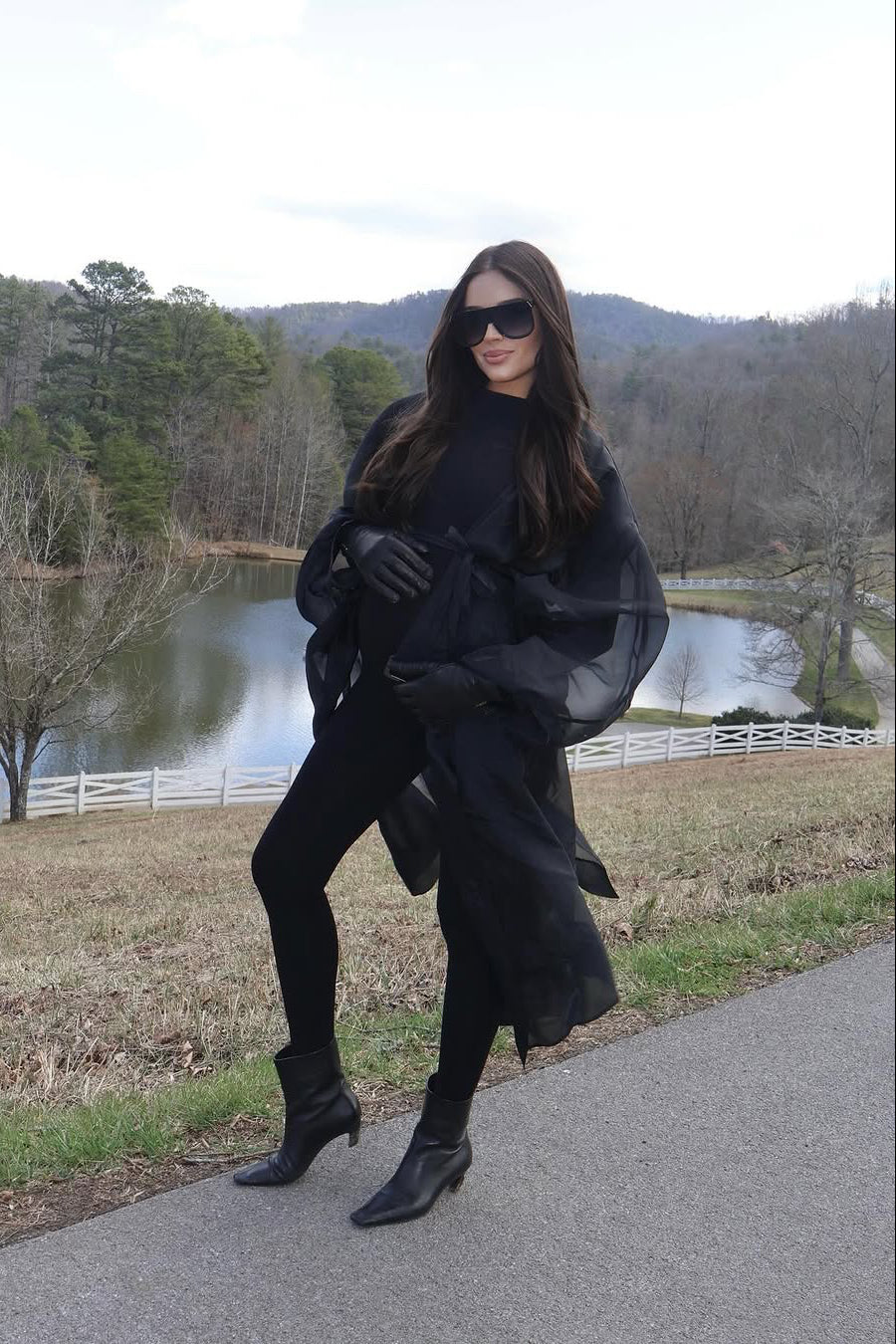 A woman posing outdoors in a black ensemble, featuring a black turtleneck jumpsuit by Bumpsuit, black gloves, and black ankle boots, set against a scenic background of a lake and wooded hills.