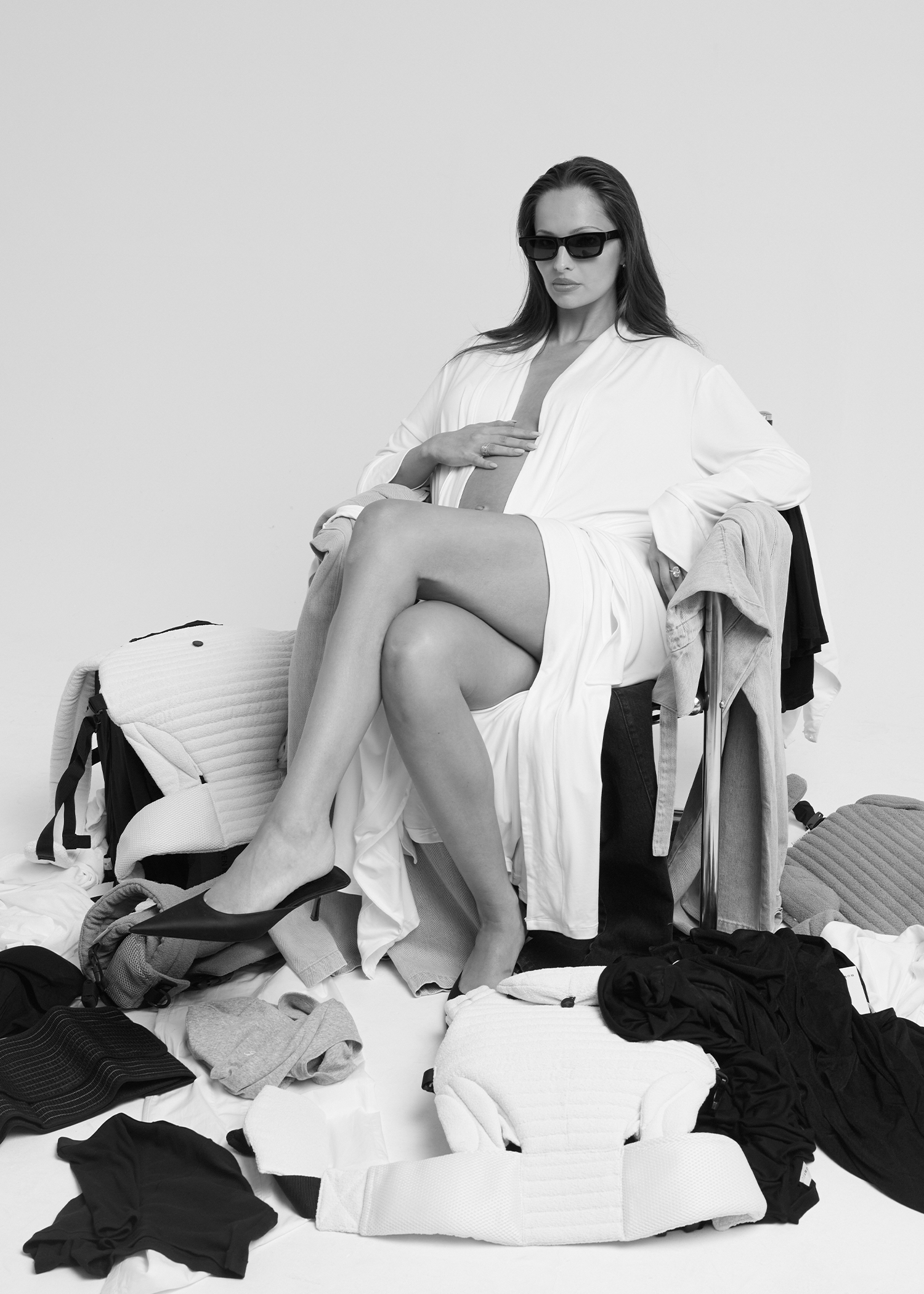 A black and white editorial photo of a pregnant model wearing sunglasses and a Bumpsuit white robe, sitting amidst piles of maternity clothing and pillows.