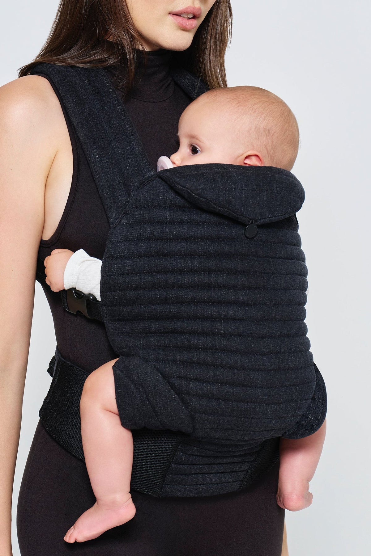 Woman holding a baby in a black  denim baby carrier by Bumpsuit against a white background