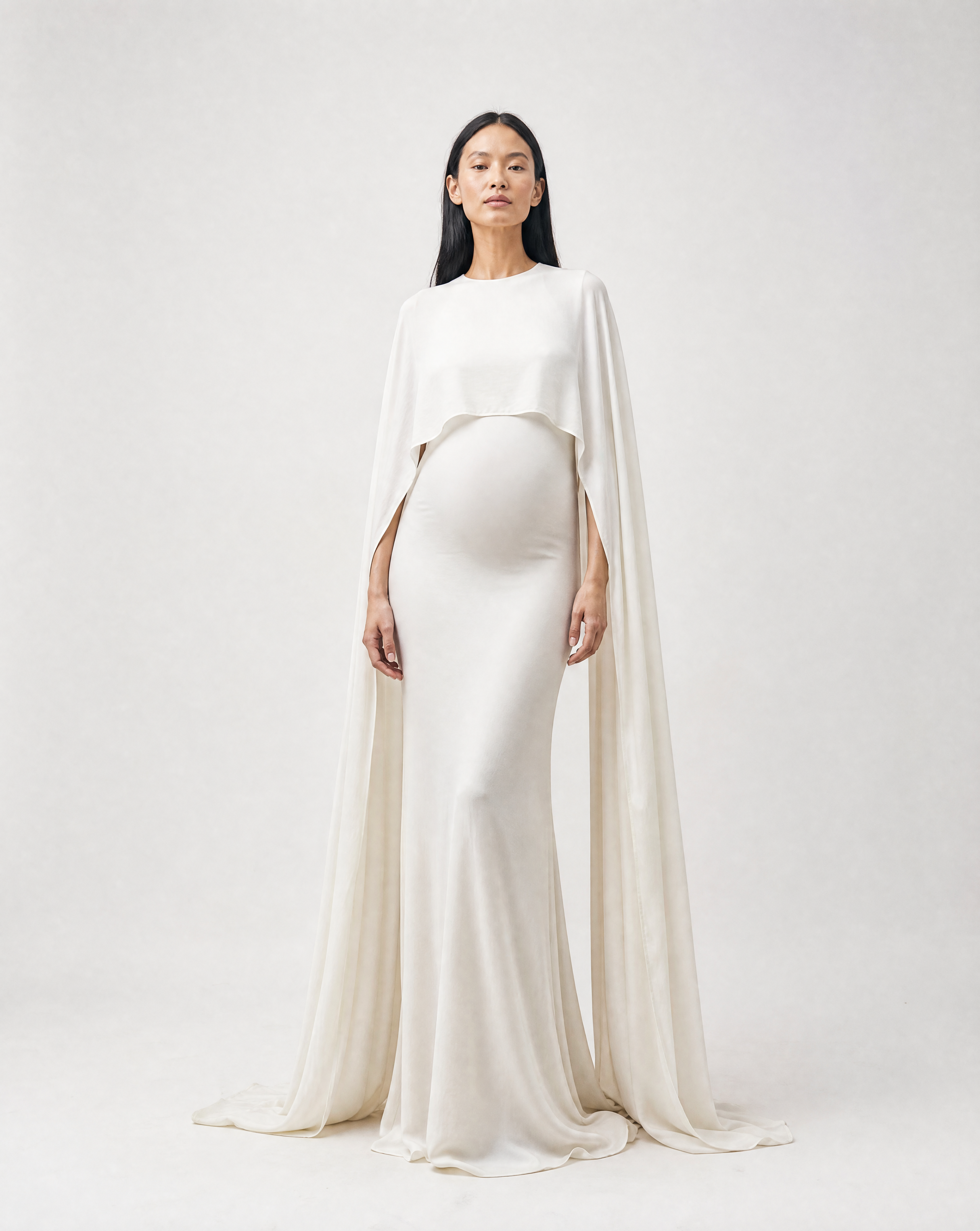 A pregnant model wearing a floor-length ivory maternity gown with a draped cape overlay, standing against a light gray studio background.