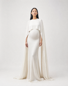 A pregnant model wearing a floor-length ivory maternity gown with a draped cape overlay, standing against a light gray studio background.