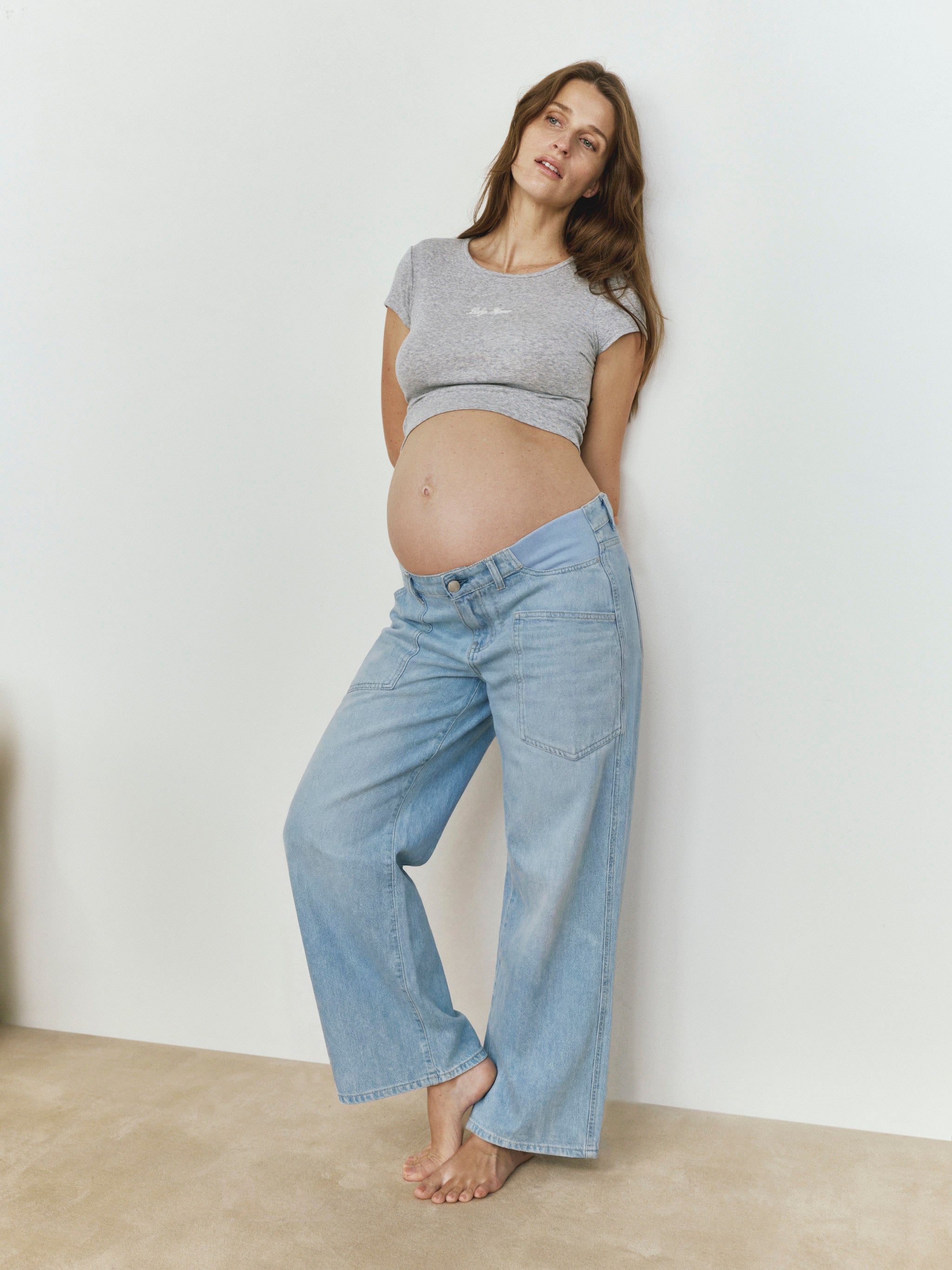 Pregnant woman wearing a Bumpsuit Heather gray baby mama tee and blue jeans against a white wall.