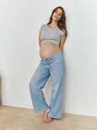 Pregnant woman wearing a Bumpsuit Heather gray baby mama tee and blue jeans against a white wall.