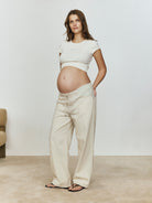 Pregnant woman wearing a white baby mama tee and beige pants against a plain background