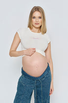 Pregnant woman wearing a white Bumpsuit  baby mama t-shirt and blue pants against a light grey background