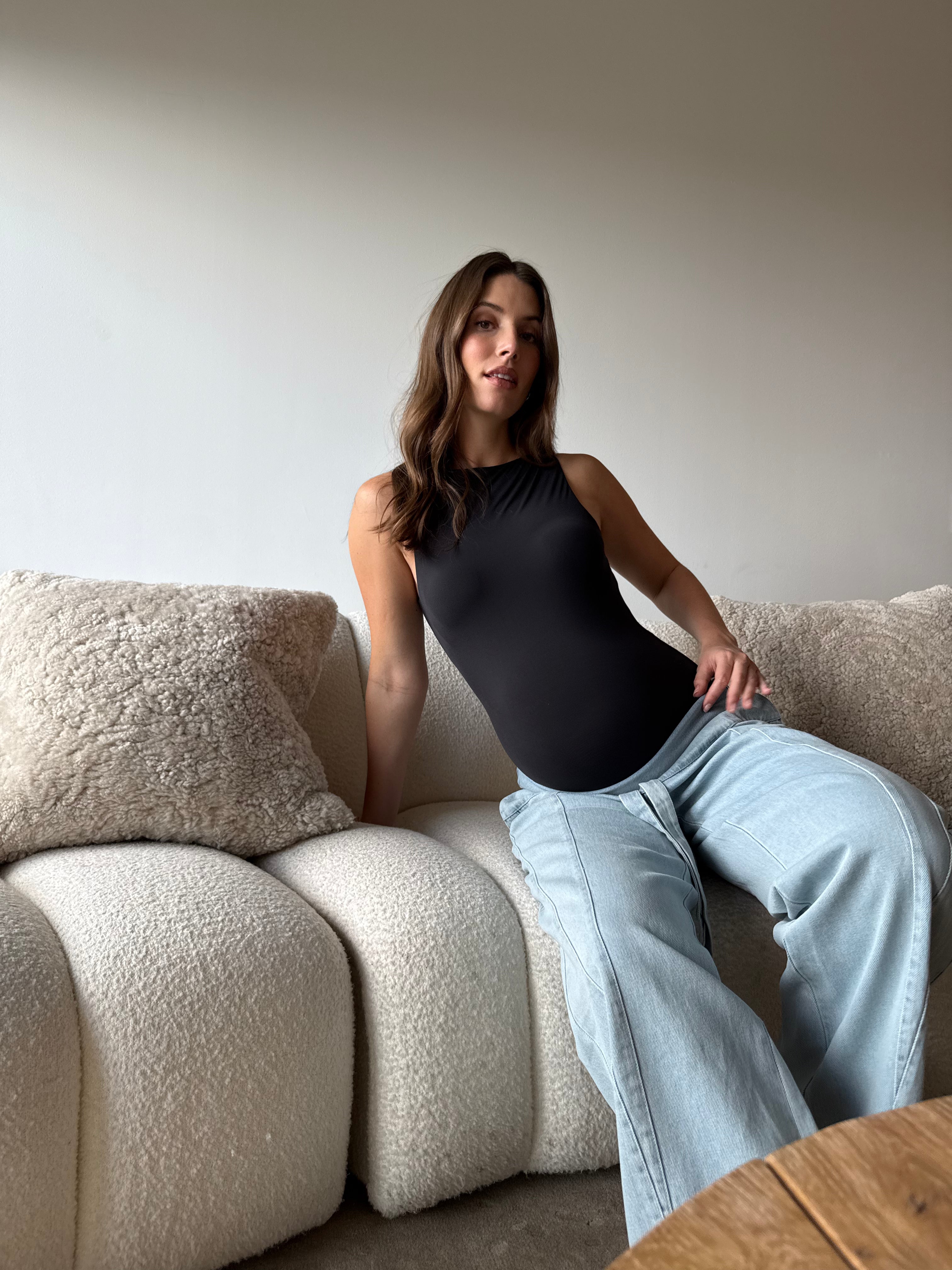 Pregnant woman wearing a black  Bumpsuit bodysuit and jeans sitting on a sofa