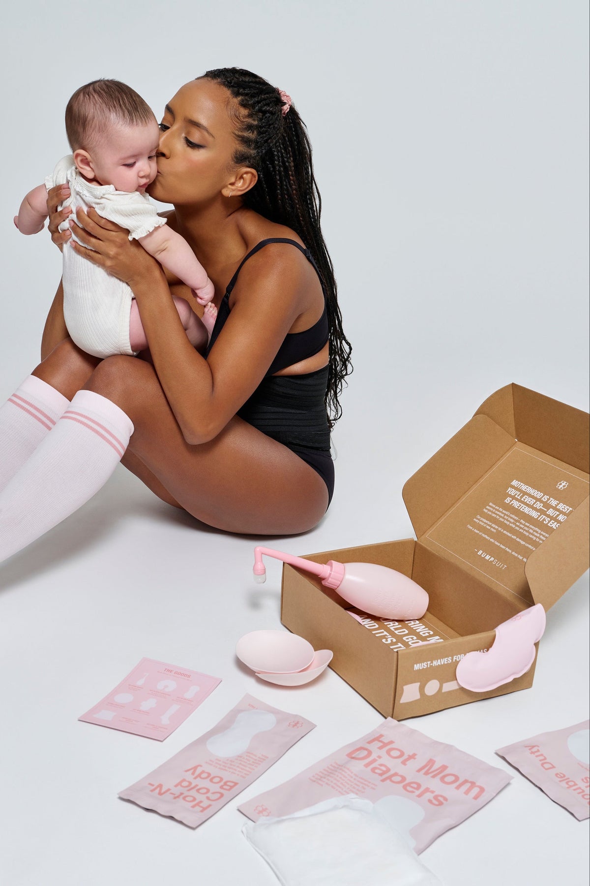 Mother kissing a baby while postpartum care kit items are arranged nearby in a studio setting