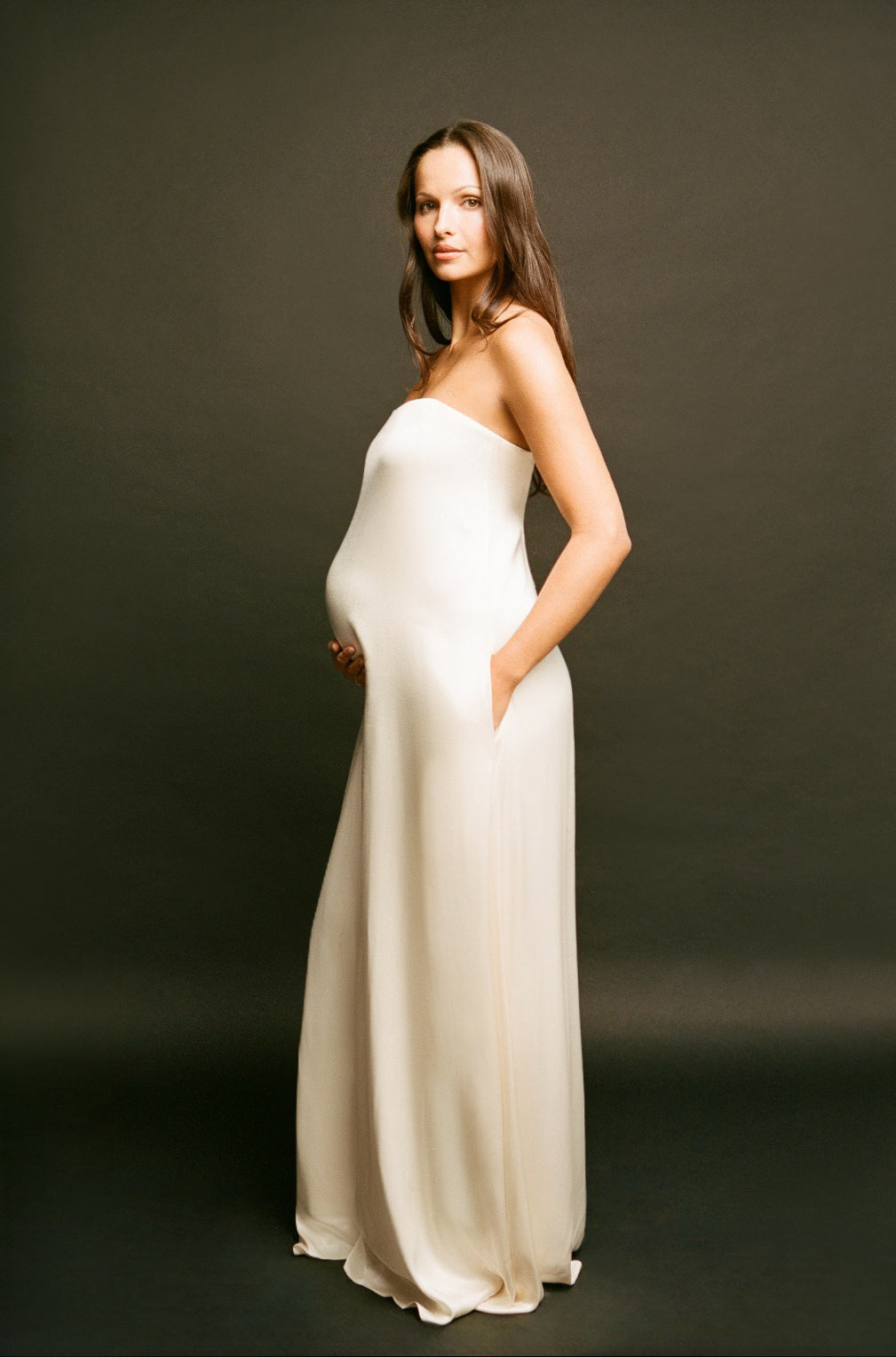 A pregnant woman wearing a Bumsuit strapless, floor-length cream satin gown stands against a dark studio background, holding her baby bump with one hand and resting the other in the side pocket of the dress.