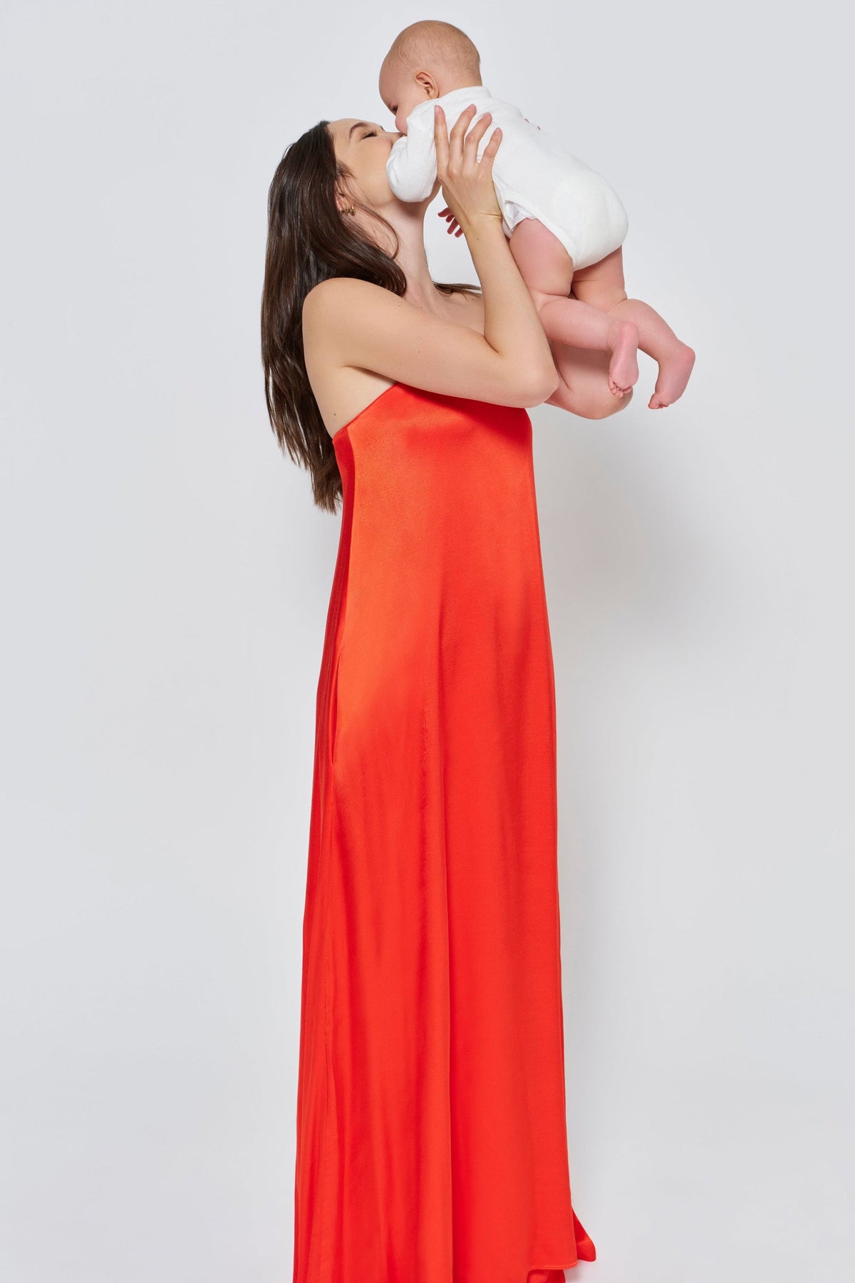 A woman wearing the Bumpsuit brushed satin strapless maxi maternity gown in red against a white background holding a baby in a white romper 