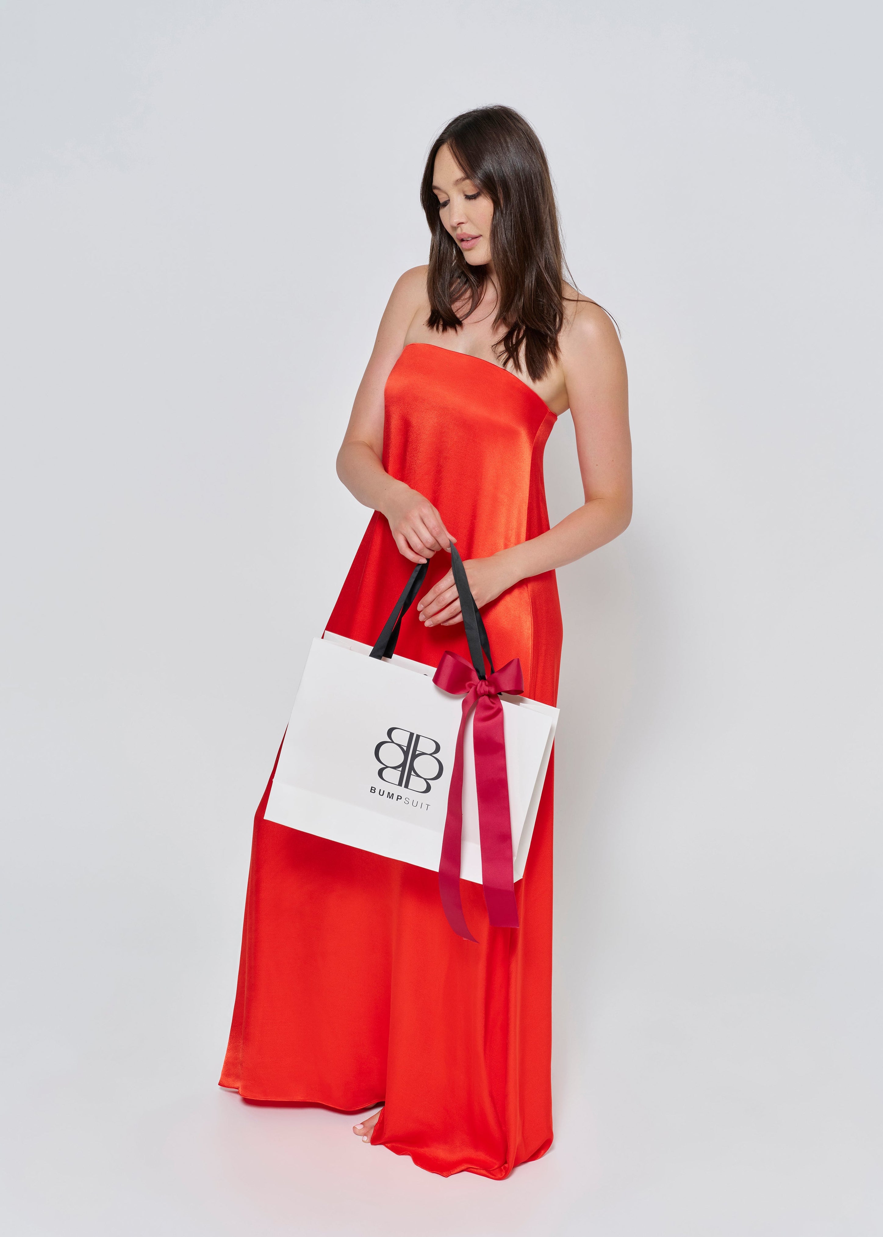 A woman wearing the Bumpsuit brushed satin strapless maxi maternity gown in red against a white background holding a Bumpsuit gift bag