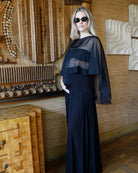A woman wearing a long black maternity dress with a chiffon cape and sunglasses, standing in a rustic room with wood-paneled walls and wooden furniture.