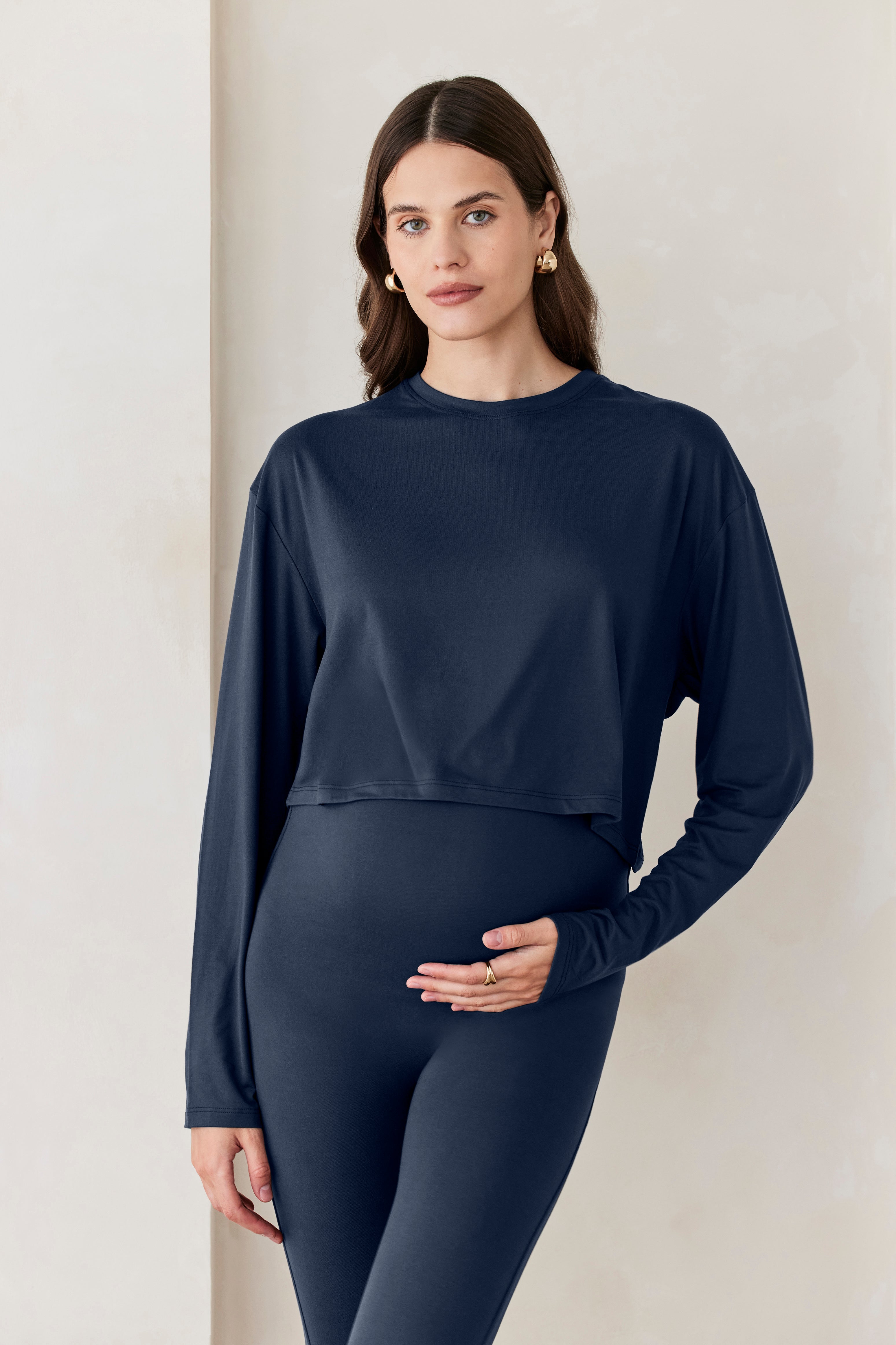 A woman standing against a beige feature wall wearing Bumpsuits Cloud  long sleeve crop tee in navy