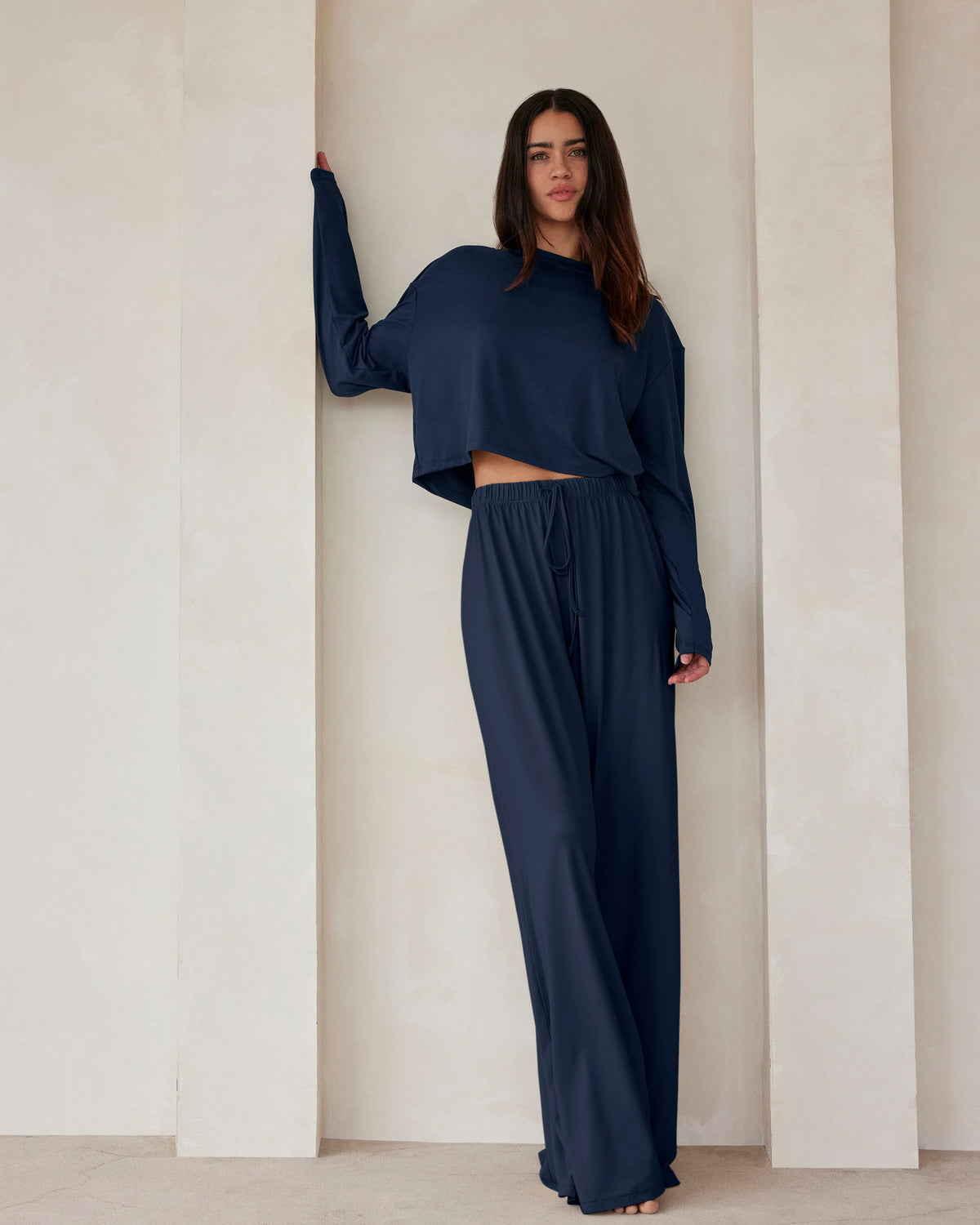 A woman standing against a beige feature wall wearing Bumpsuits Cloud lounge pants and a long sleeve crop tee in navy