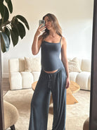 Pregnant woman taking a mirror selfie wearing a Bumpsuit cloud cement blue tank top and pants in a living room.