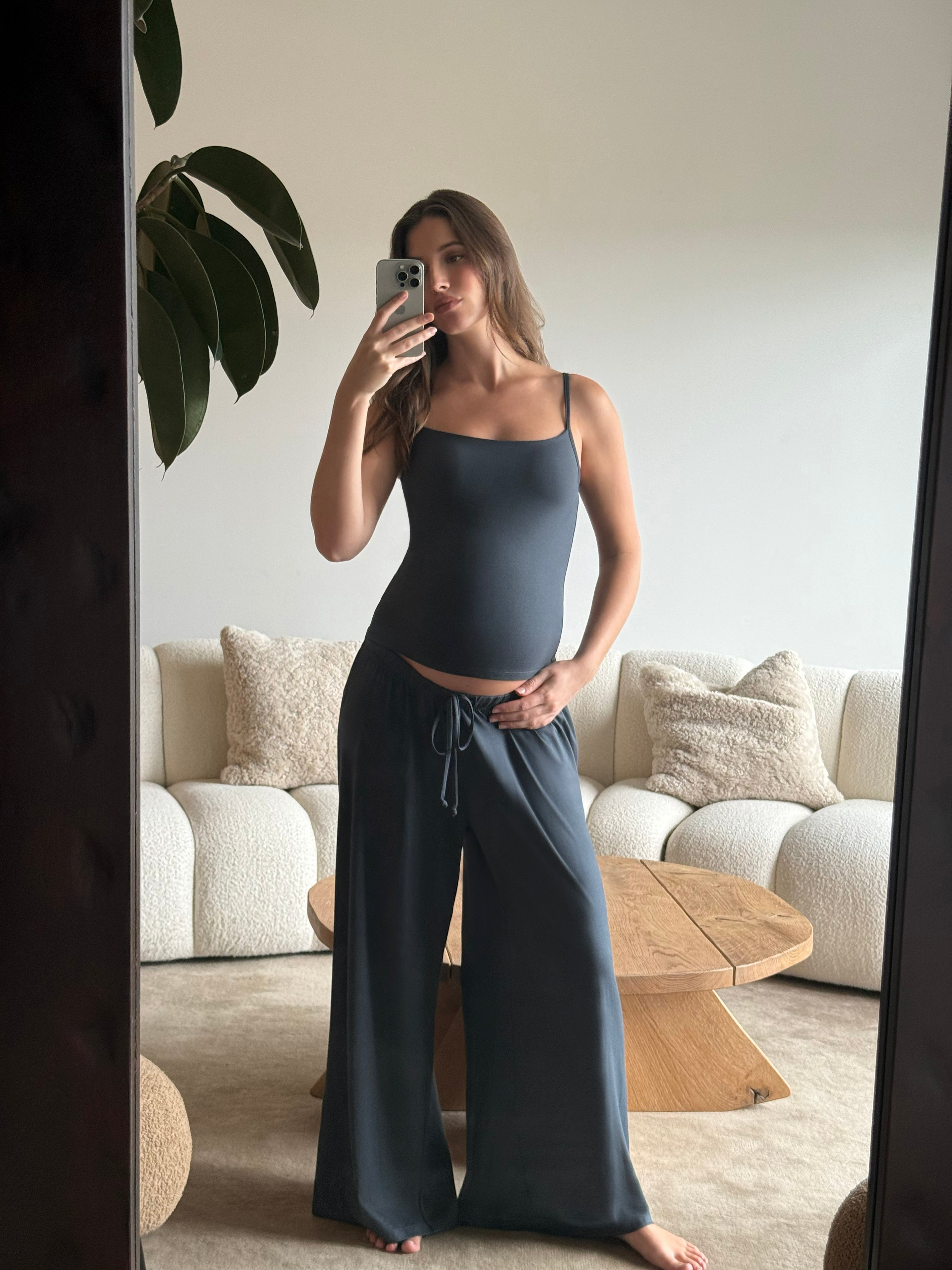 Pregnant woman taking a mirror selfie wearing a Bumpsuit cloud cement blue tank top and pants in a living room.