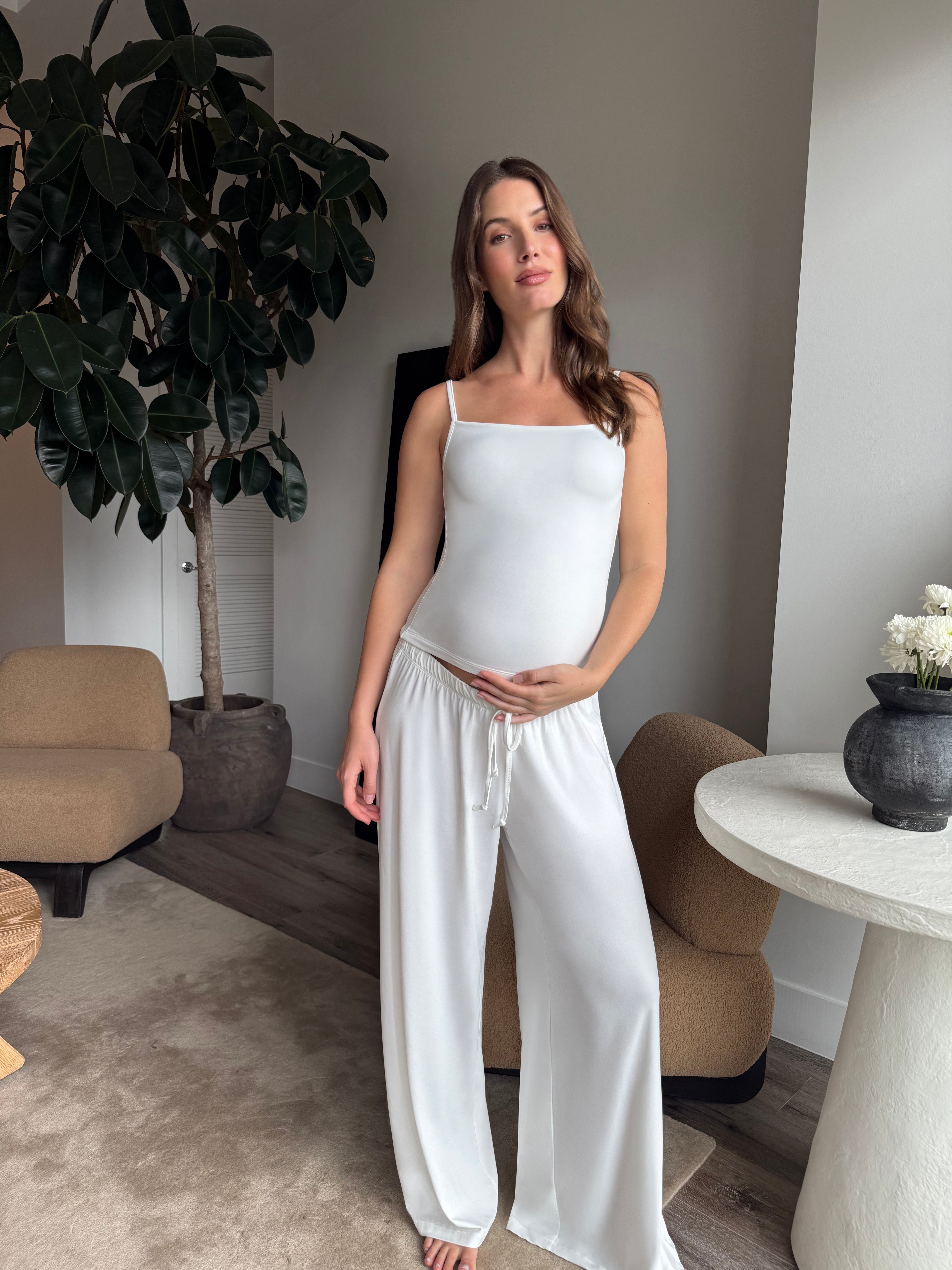 Pregnant woman wearing a Bumpsuit cloud ice pink tank top and pants in a living room.