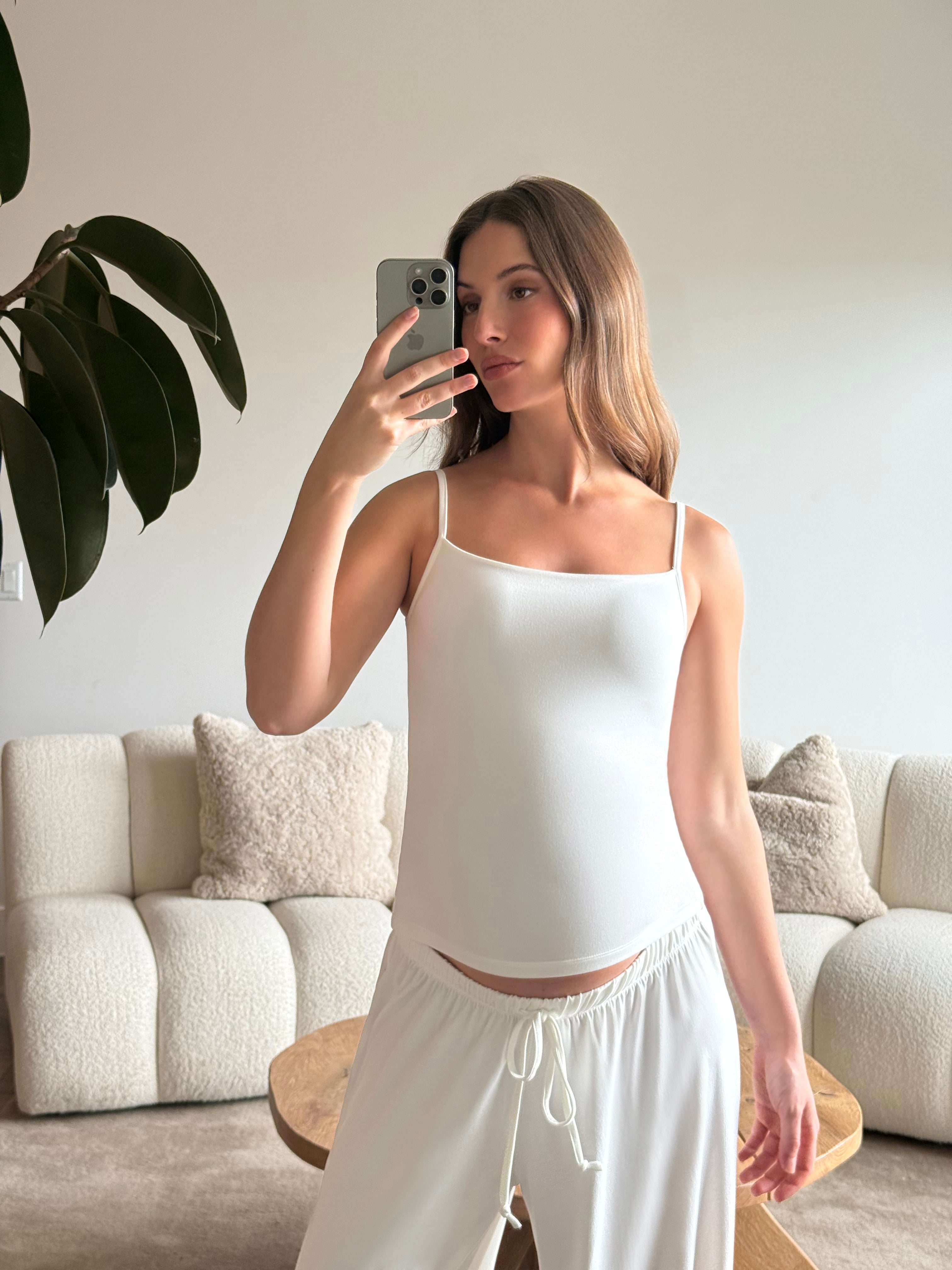 Pregnant woman taking a mirror selfie wearing a Bumpsuit cloud ivory tank top and pants in a living room.