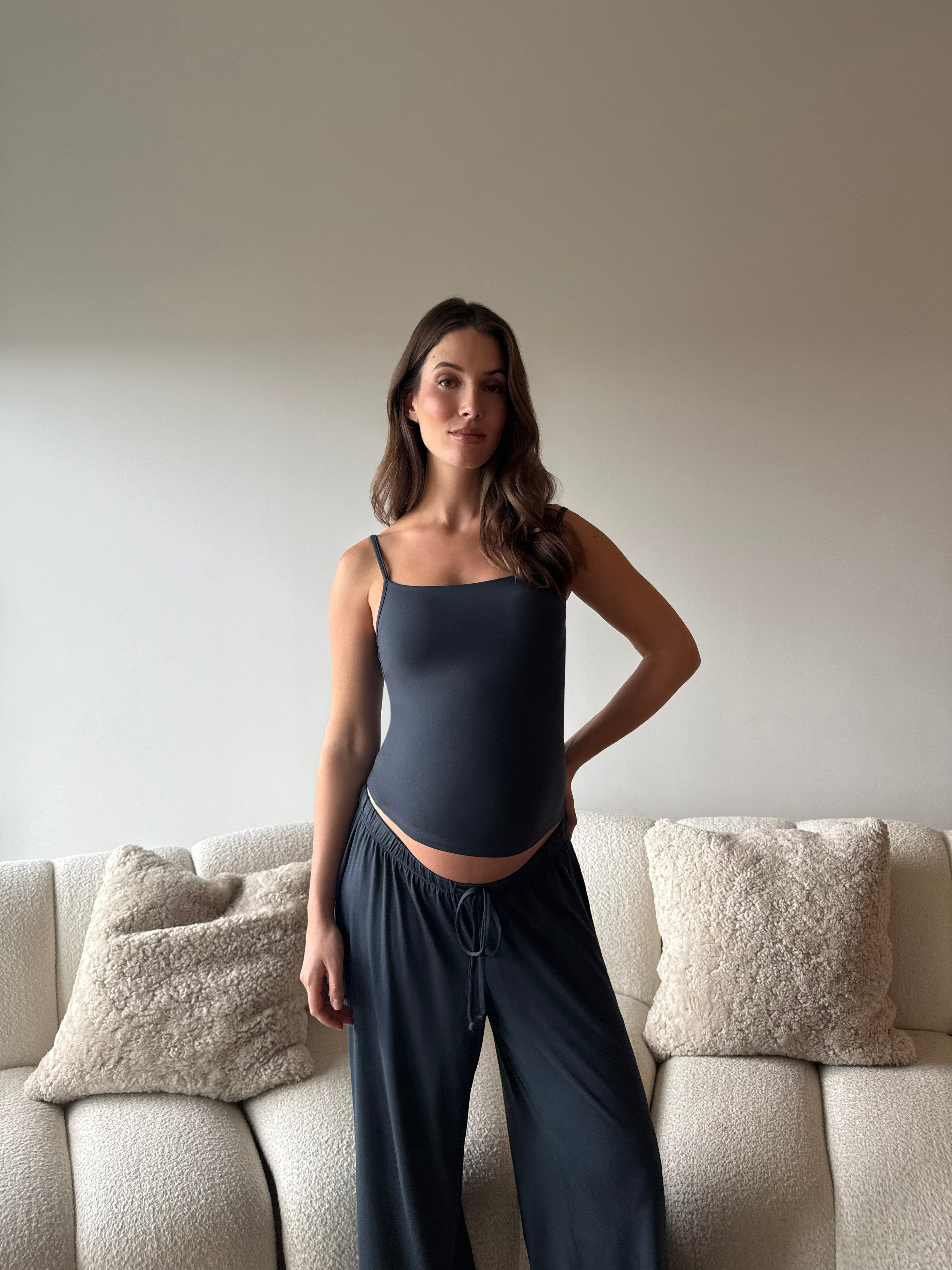 Pregnant woman wearing a Bumpsuit cloud cement blue tank top and pants in a living room.