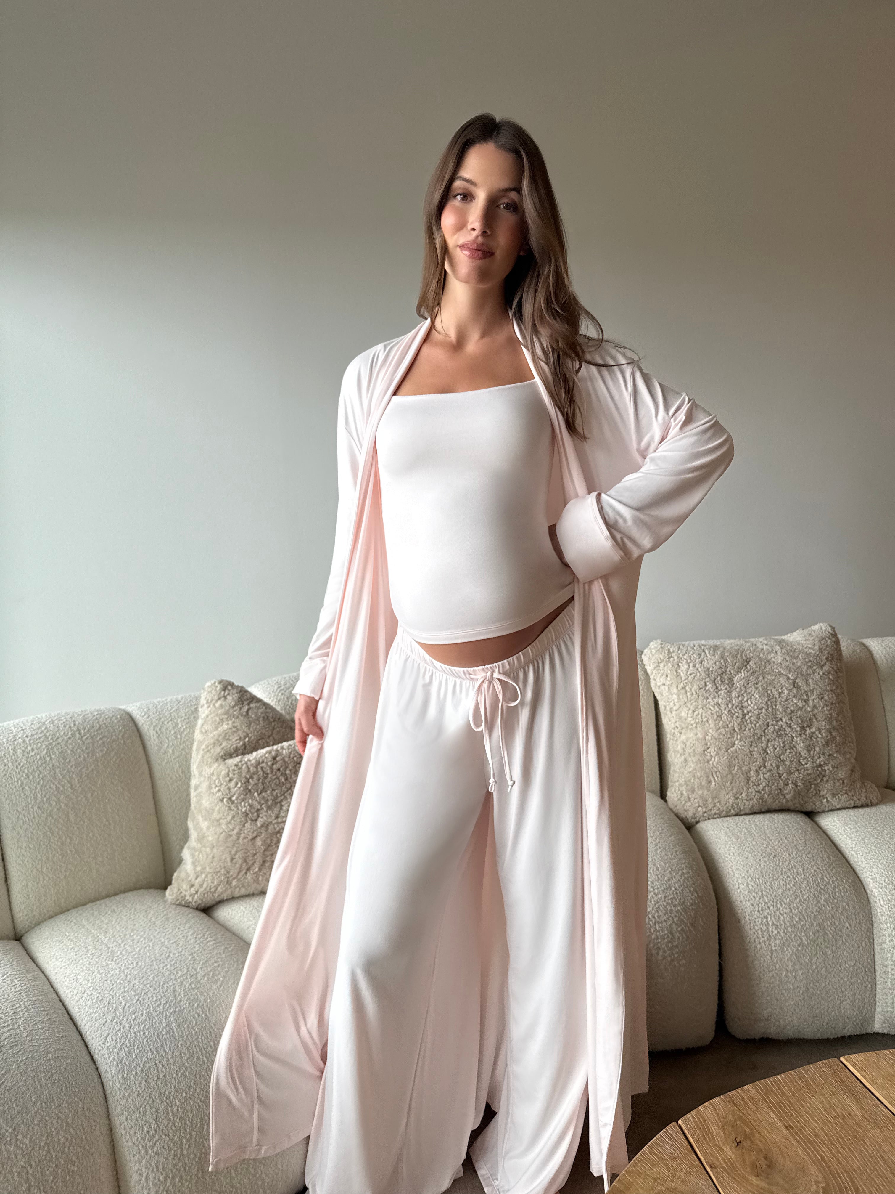 Pregnant woman taking a mirror selfie wearing a Bumpsuit cloud ice pink tank top, robe and pants in a living room.