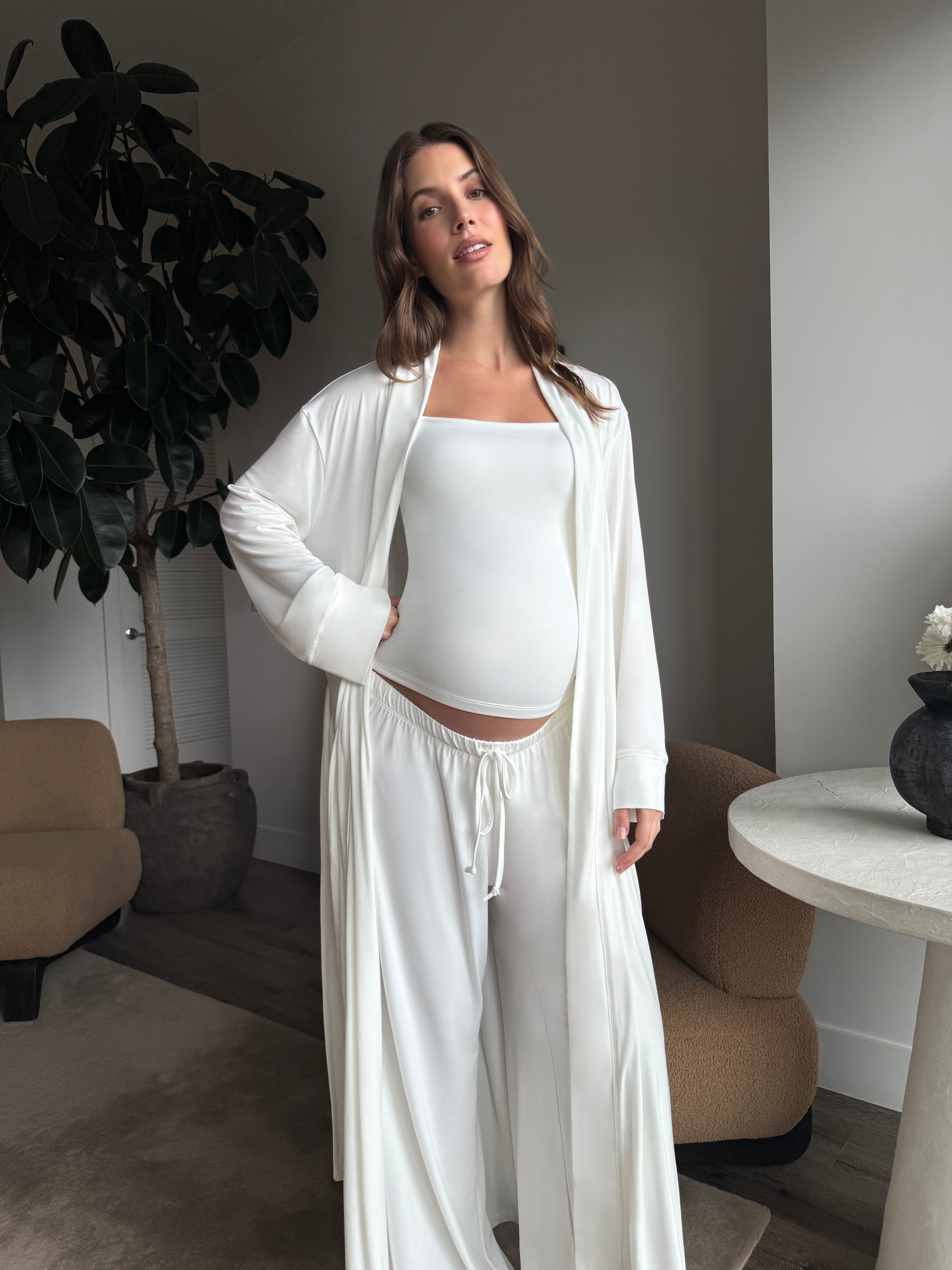 Pregnant woman wearing a Bumpsuit cloud ivory tank top, robe and pants in a living room.