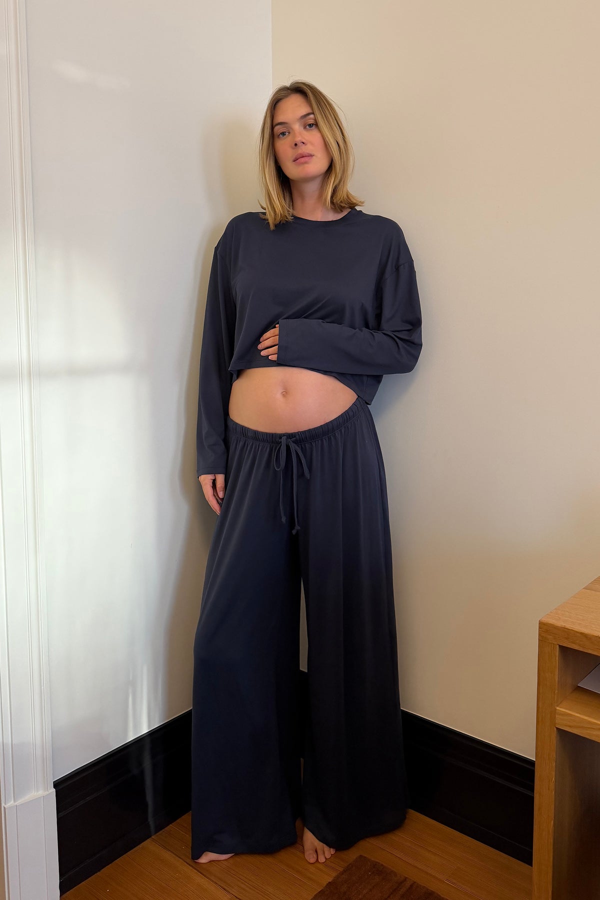 A woman modeling a matching navy blue loungewear set consisting of a long-sleeve cropped top and wide-leg drawstring pants, standing in a corner of a room with beige walls and wooden flooring.