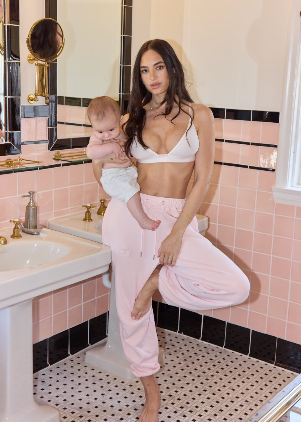 Woman holding a baby in a bathroom with pink tiles and a sink.