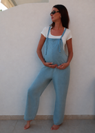 A pregnant woman wearing Bumsuits light blue denim overalls over a white t-shirt, posing against a white stucco wall.
