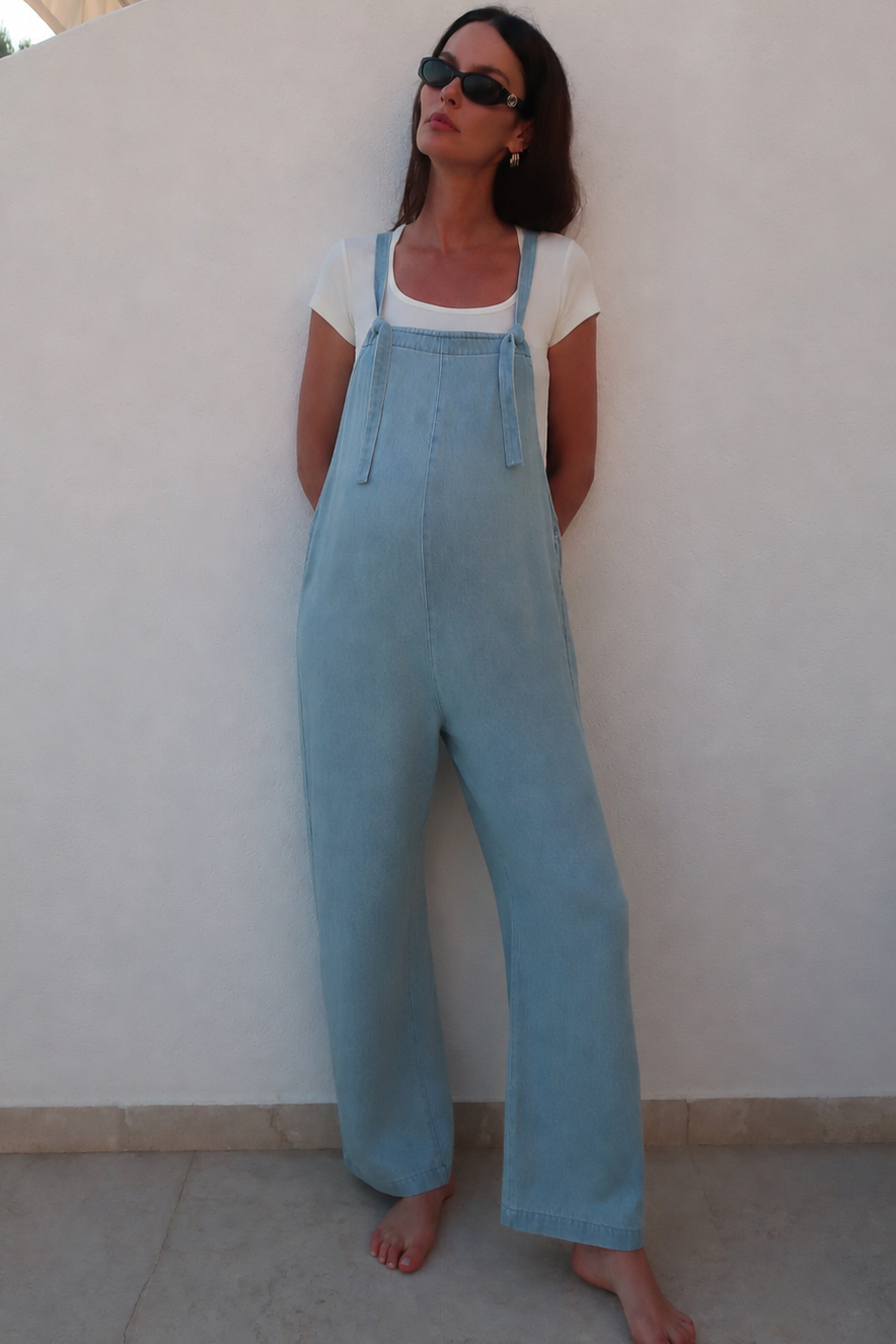 A woman wearing a light blue denim overall jumpsuit over a white t-shirt, standing barefoot against a white wall.