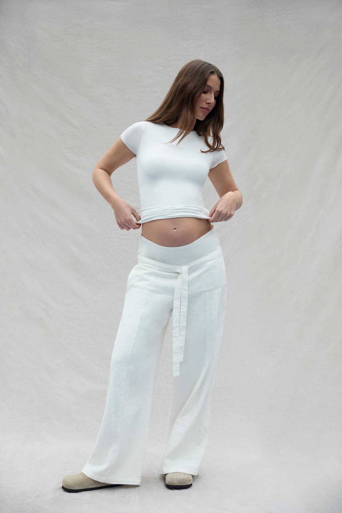 Woman wearing a white Bumpsuit shit and relaxed fisherman pants against a plain background