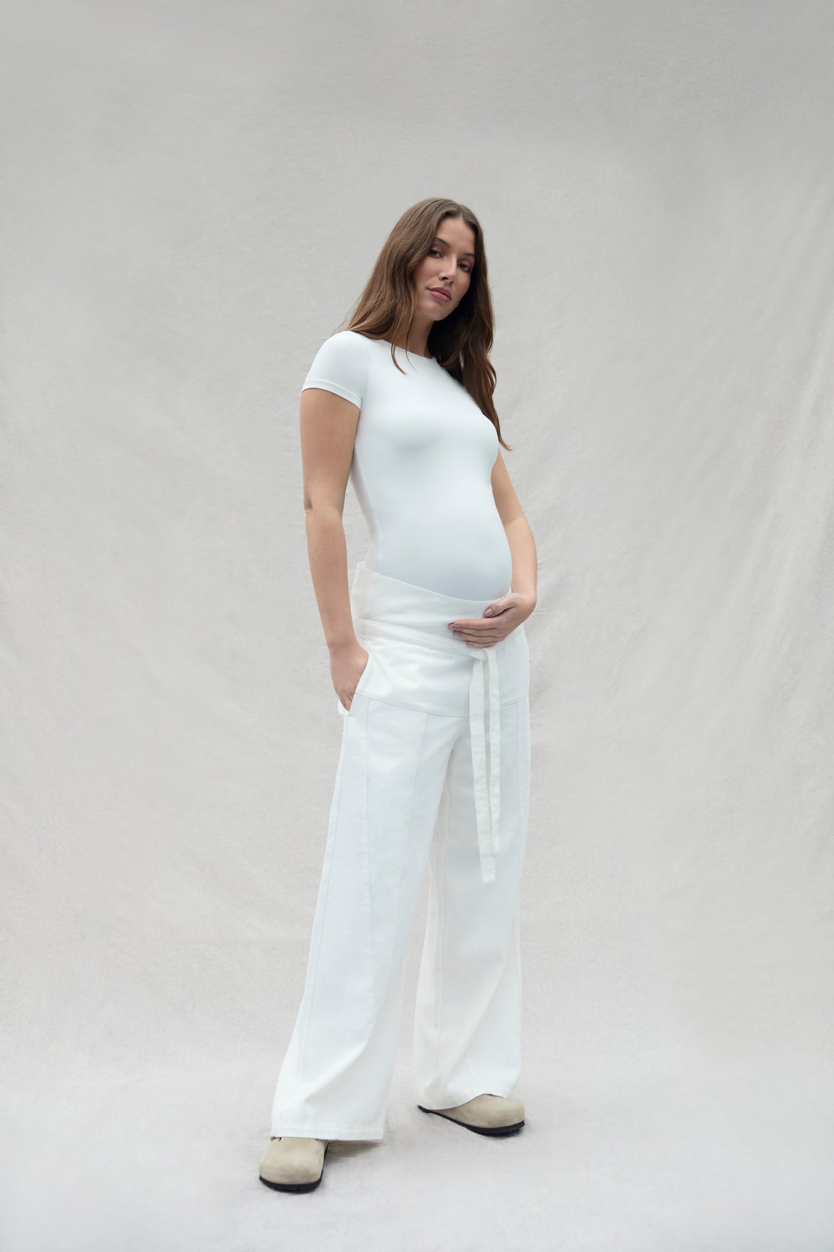 Pregnant woman wearing a white Bumpsuit outfit against a plain background