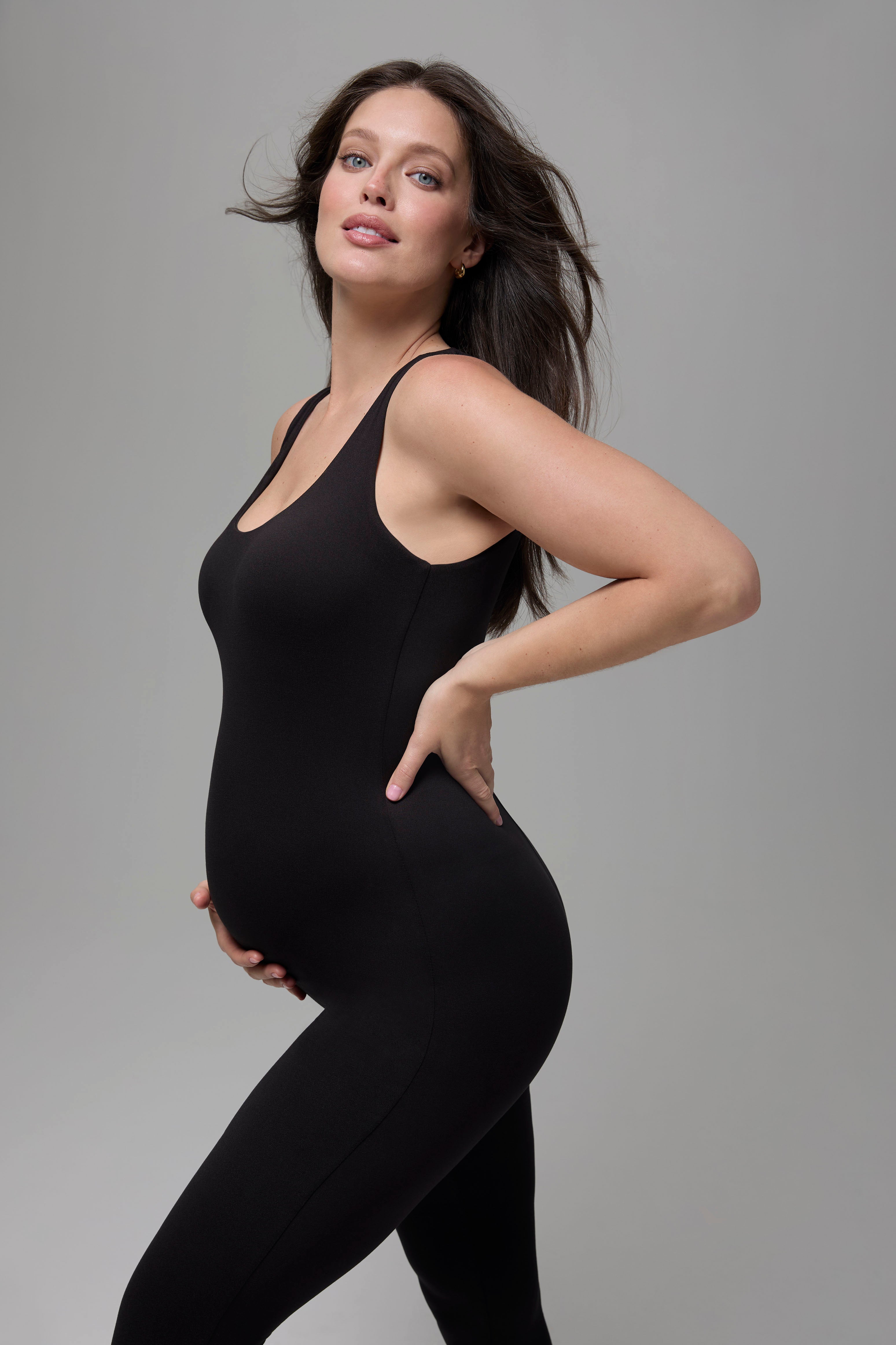 Pregnant woman wearing a black Bumpsuit bodysuit against a grey background