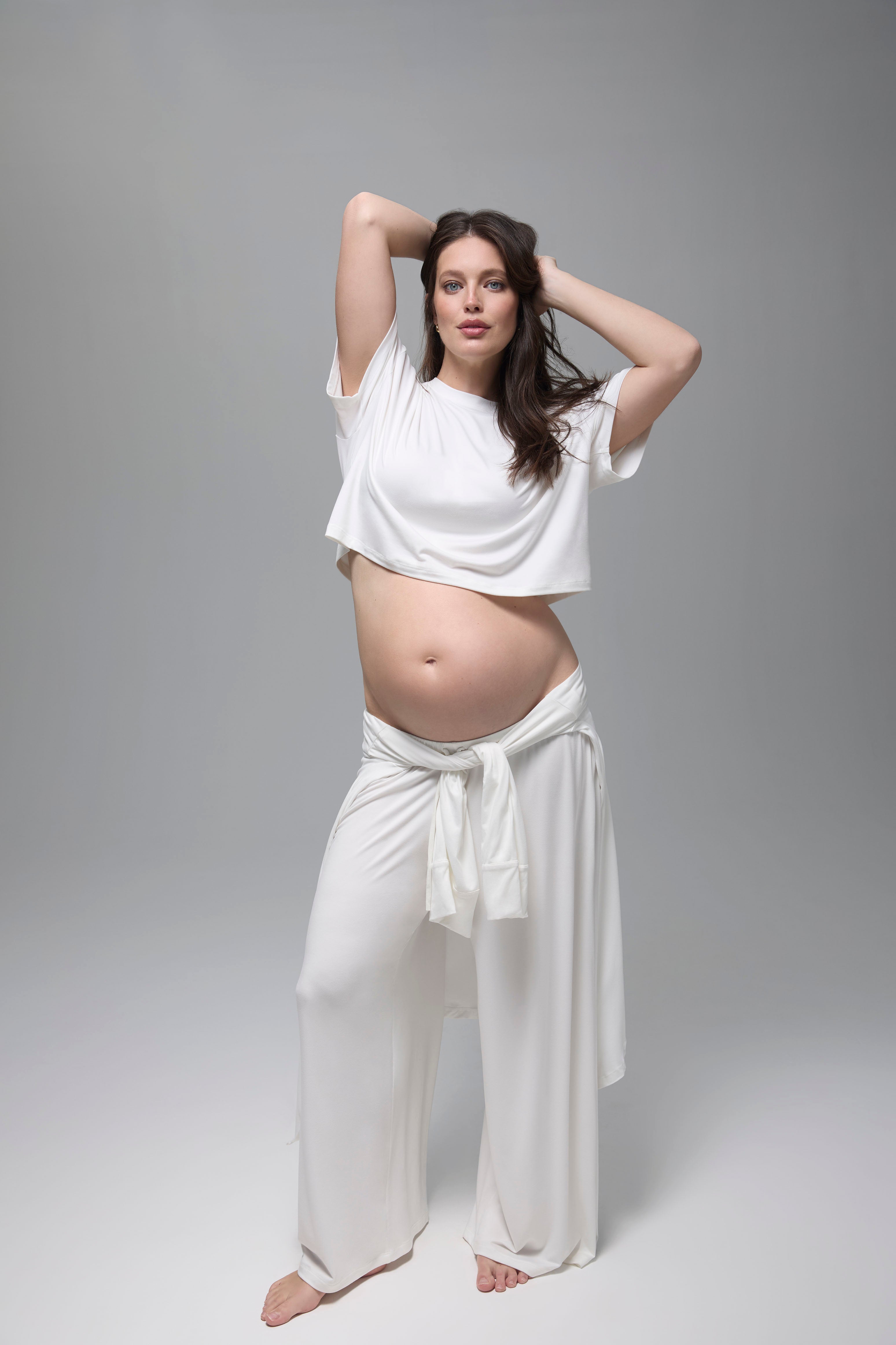 Pregnant woman wearing a Bumpsuit white night lounge outfit against a grey background