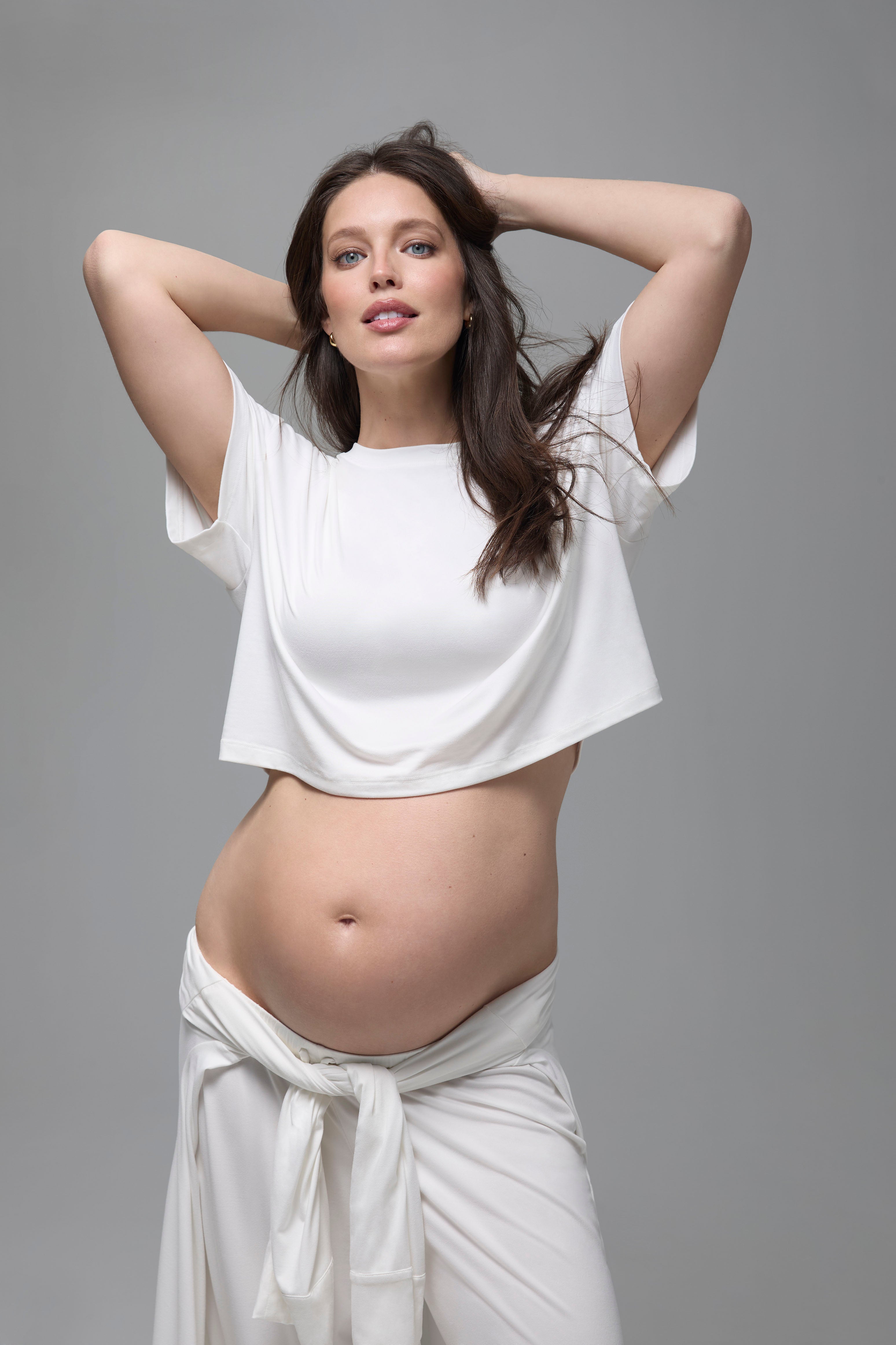 Pregnant woman wearing a white bumsuit outfit against a gray background