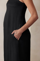 A pregnant woman wearing the Bumpsuit brushed satin strapless maxi maternity gown in black against a beige feature wall with hands in her pockets