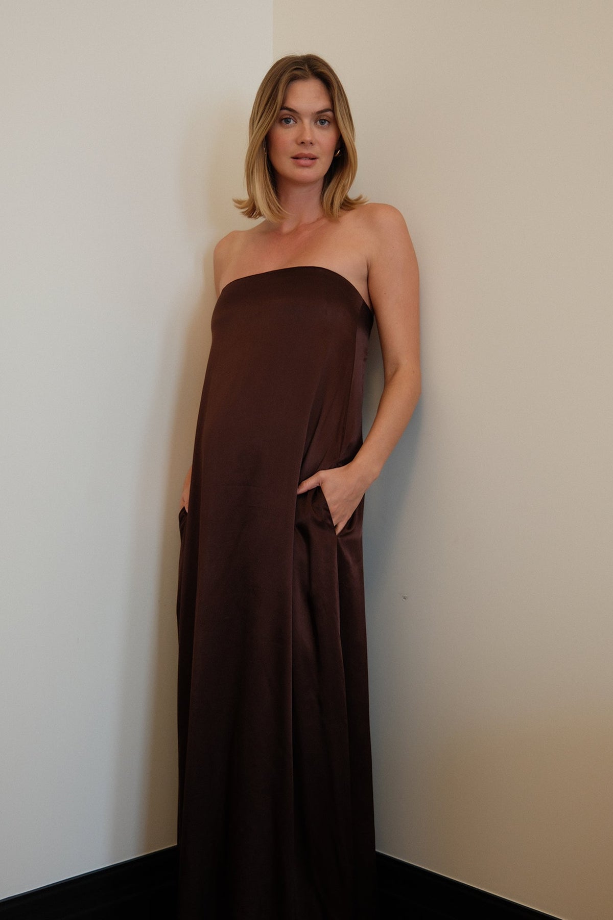 Pregnant woman standing in the corner of a room with her hands in her pockets, wearing a chocolate brown, brushed satin, strapless Bumpsuit maternity gown.