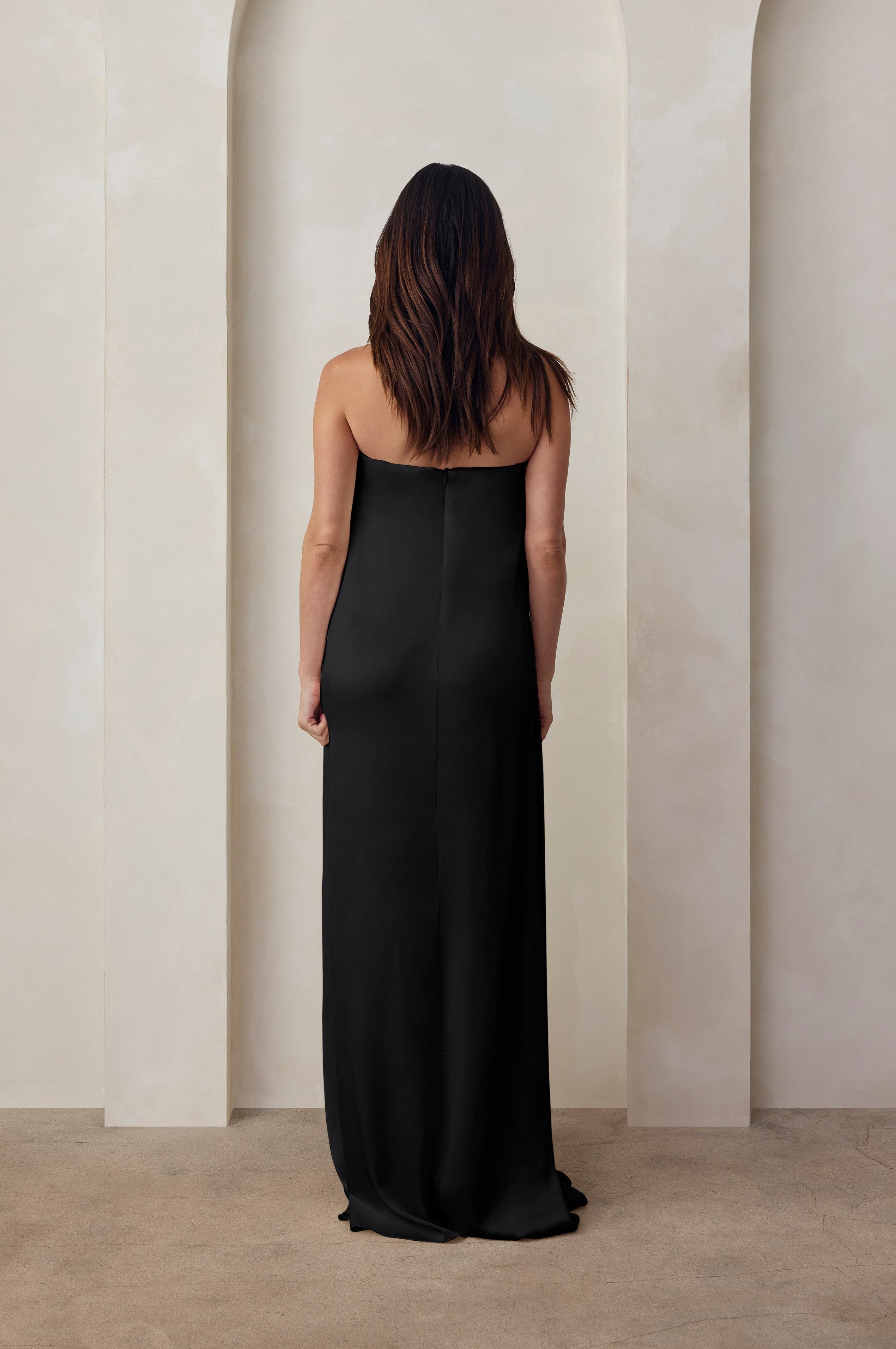 Back view of a pregnant woman wearing the Bumpsuit brushed satin strapless maxi maternity gown in black against a beige feature wall