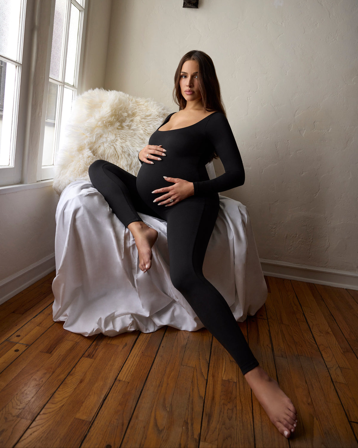 A pregnant woman wearing a black long-sleeve jumpsuit sits barefoot on a draped white chair in a room with hardwood floors and a window.