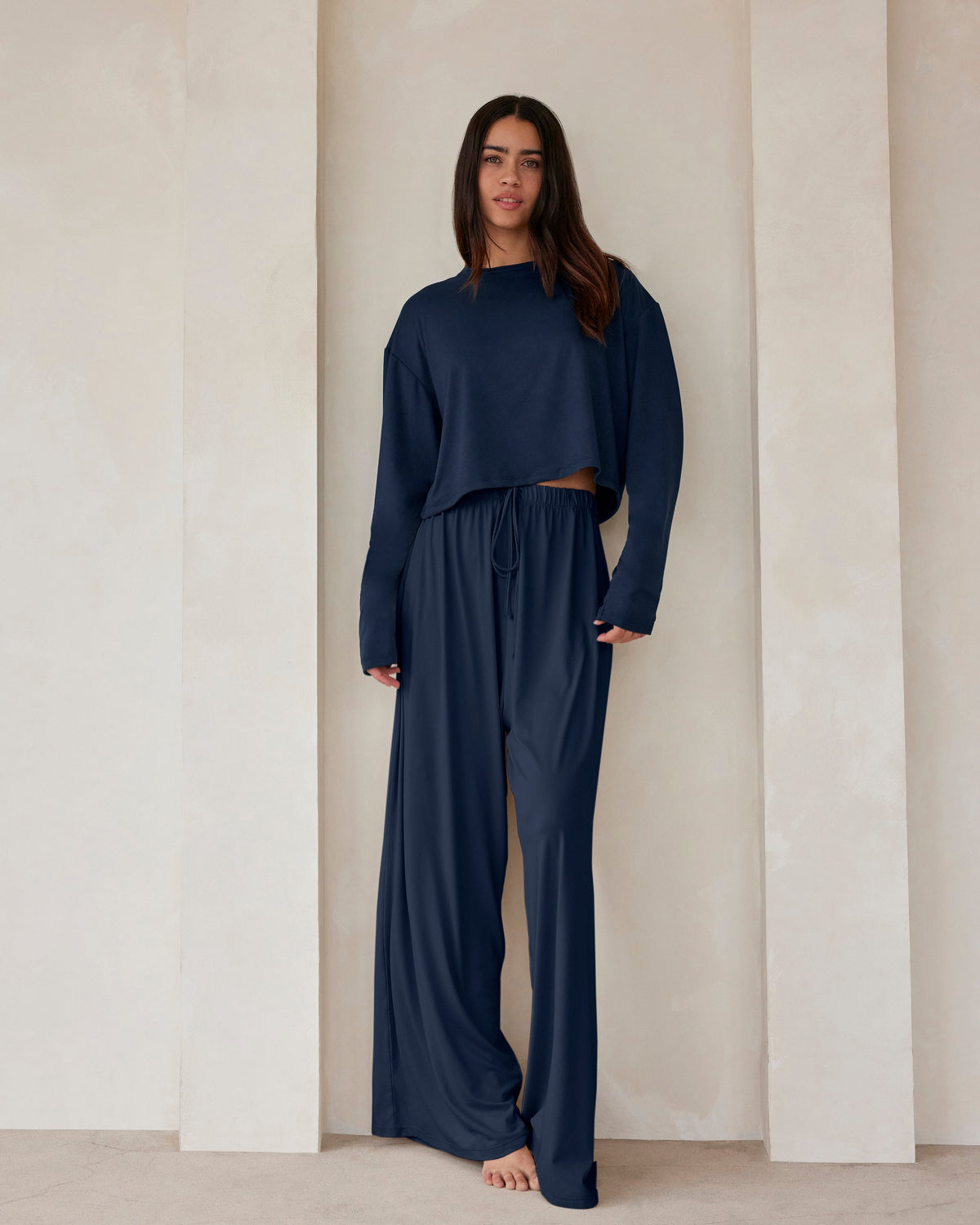 A woman standing against a beige wall in navy lounge pants and a crop long sleeve tee by Bumpsuit